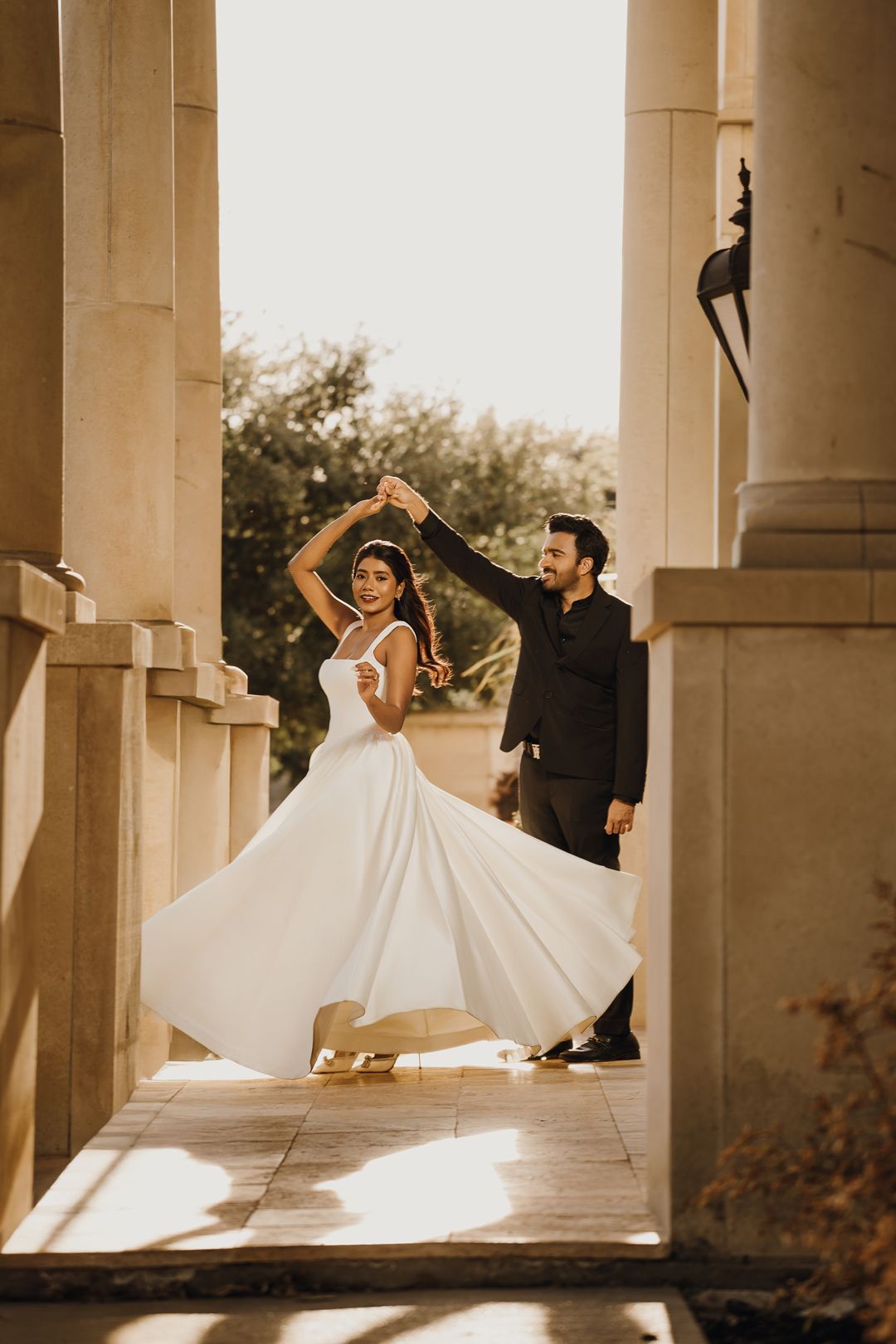 Couple dancing elegantly between stone columns, bride in flowing white gown twirling as groom in black suit leads her.