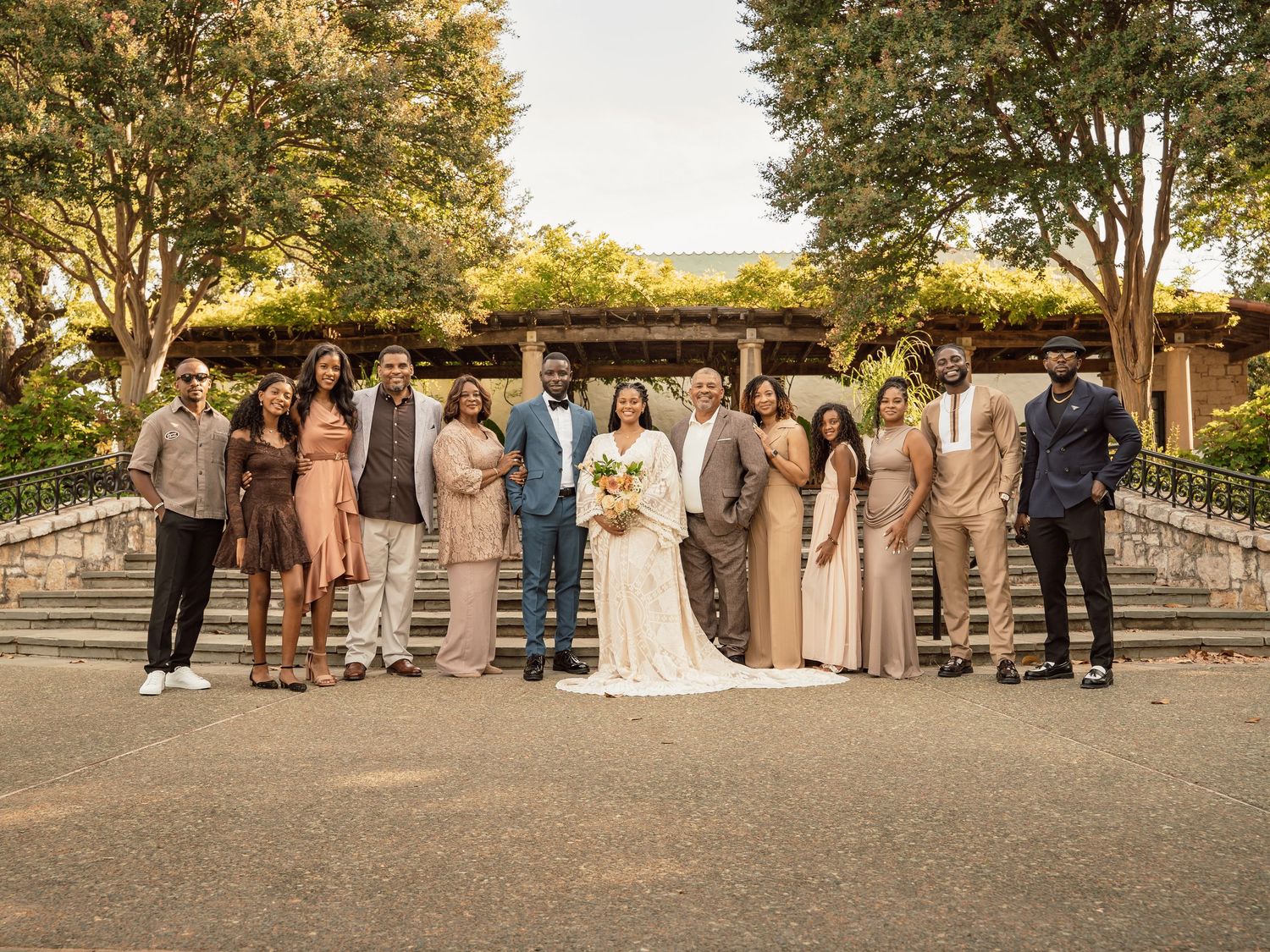 Wedding party poses outdoors in neutral tones, bride in white gown, groom in blue suit, trees and pavilion behind.