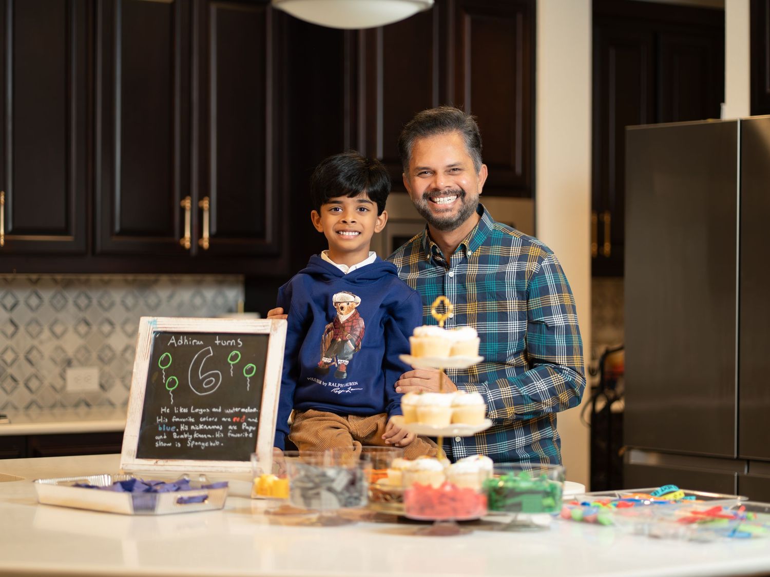 A colorful tiered birthday cake sits on a kitchen counter with dark cabinets and decorative elements in the background.