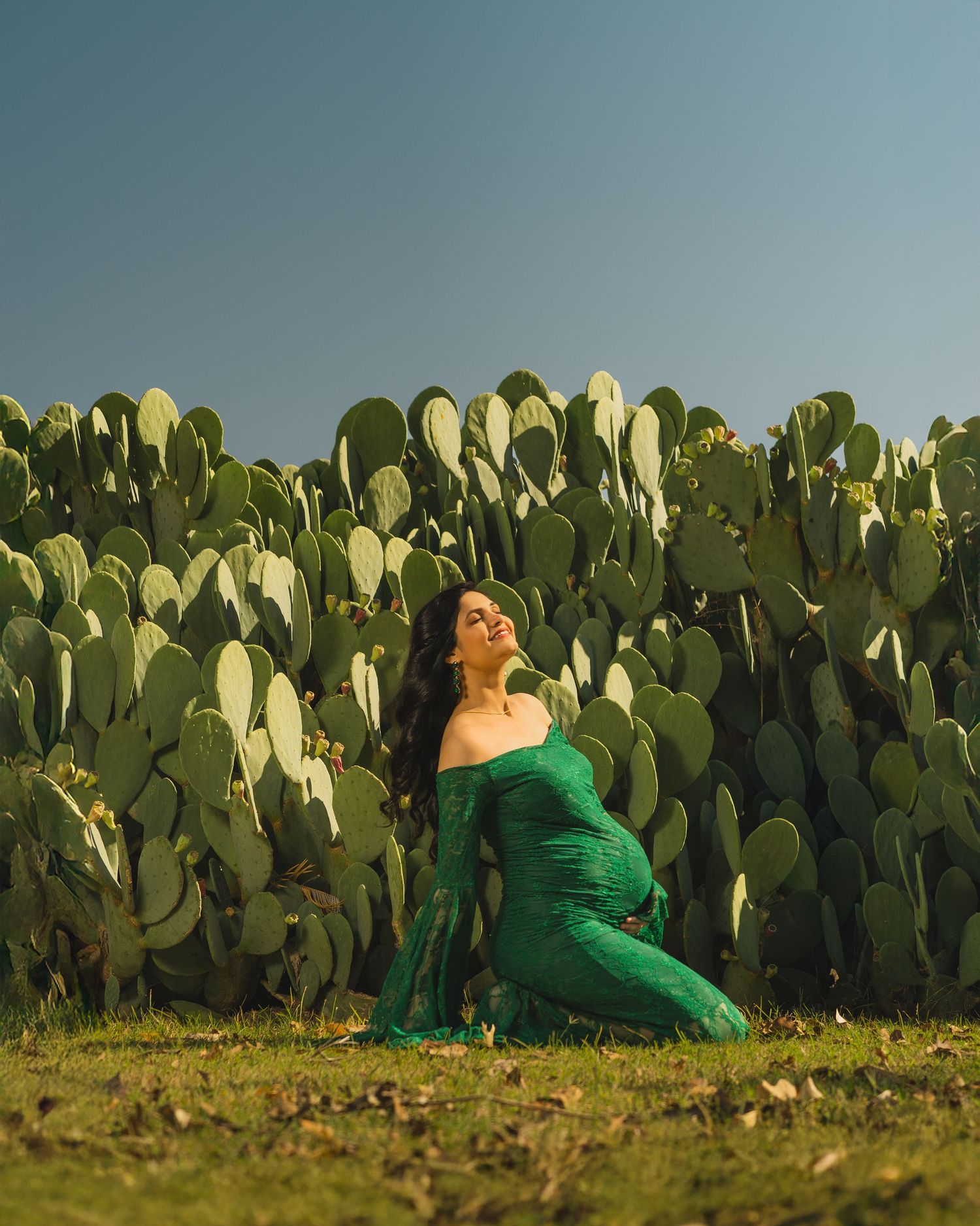 Pregnant woman in green dress poses gracefully before large prickly pear cacti under clear blue sky.