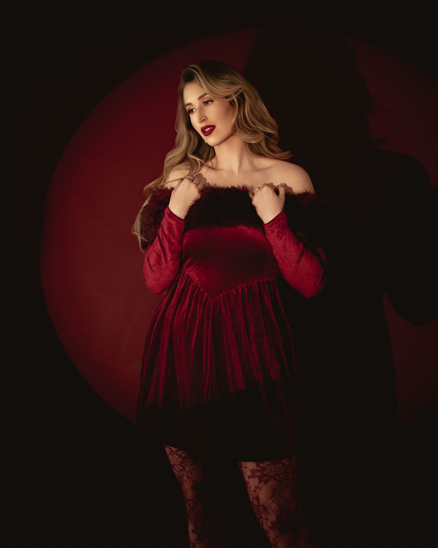 Woman in red velvet off-shoulder mini dress with lace tights poses dramatically against dark red background.