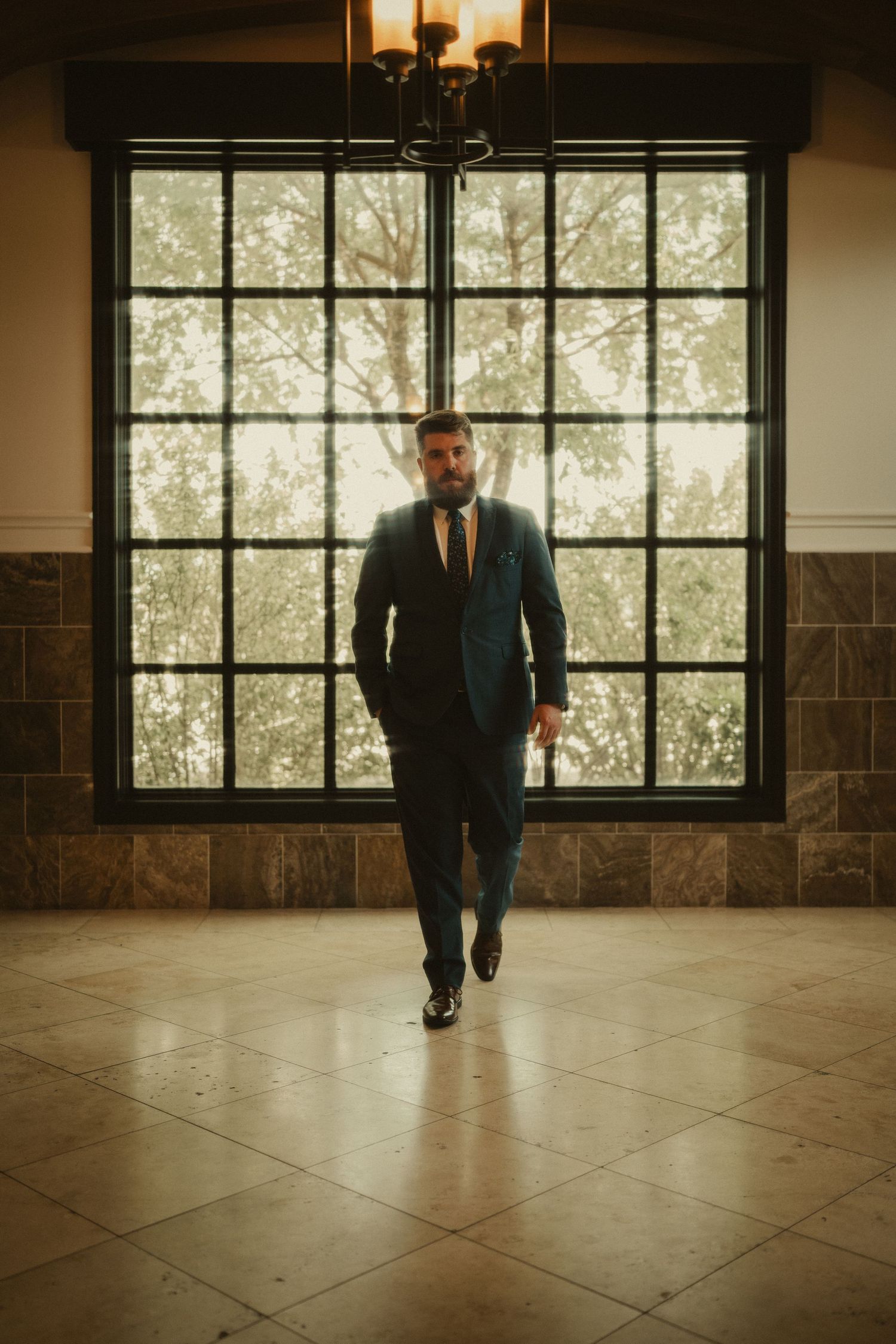 A bearded man in a navy blue suit walks confidently toward the camera in an elegant tiled room with large windows.