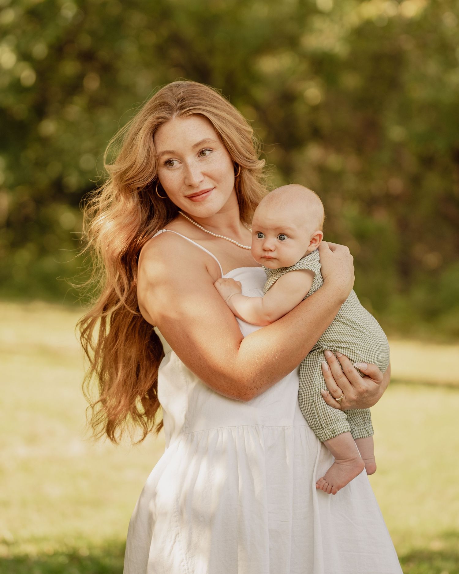 Mother in white dress twirling and dancing with baby in outdoor garden setting during golden hour.