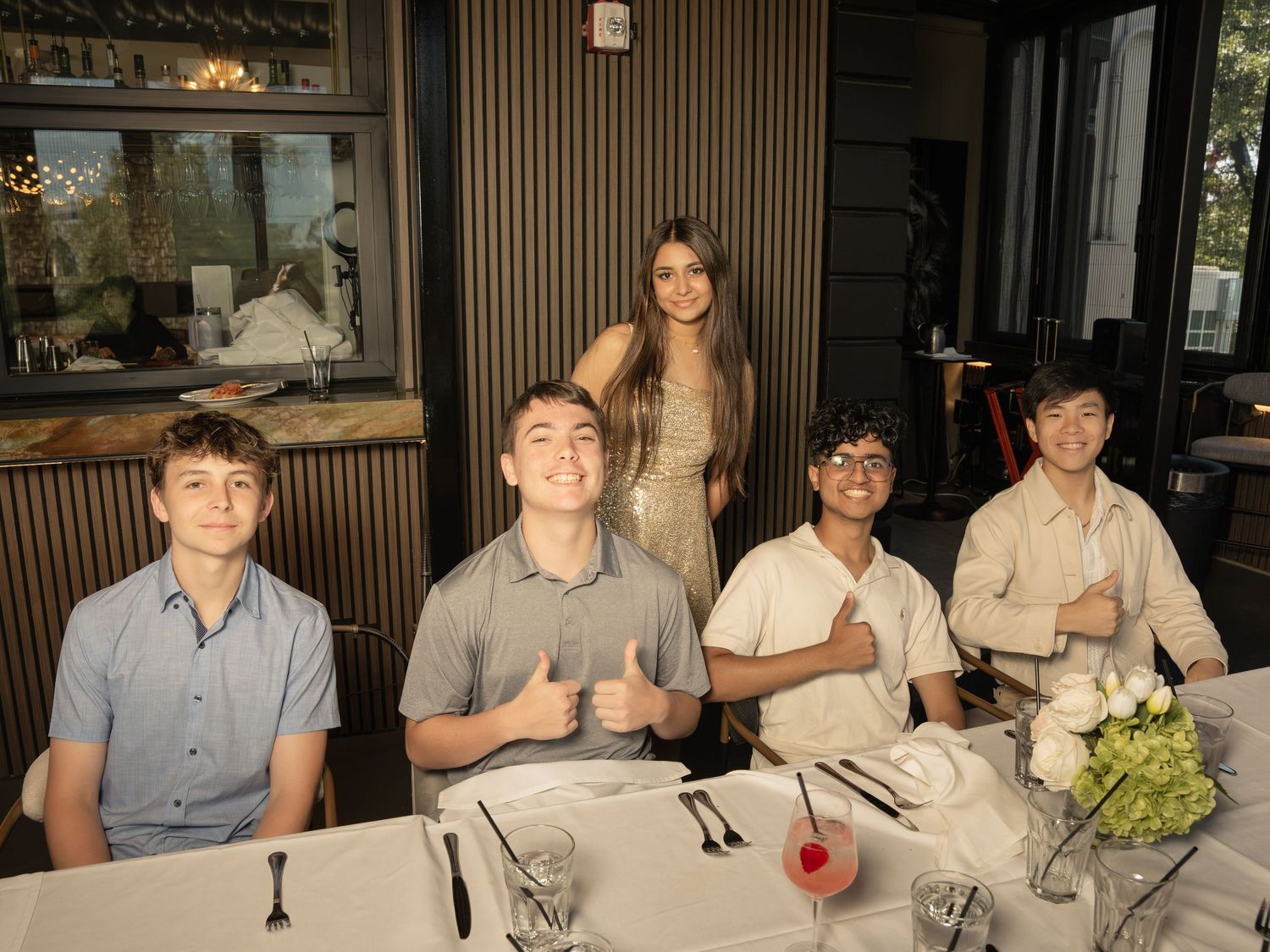 Five teenagers posing at a restaurant table, four boys seated giving thumbs up and a girl standing behind them.