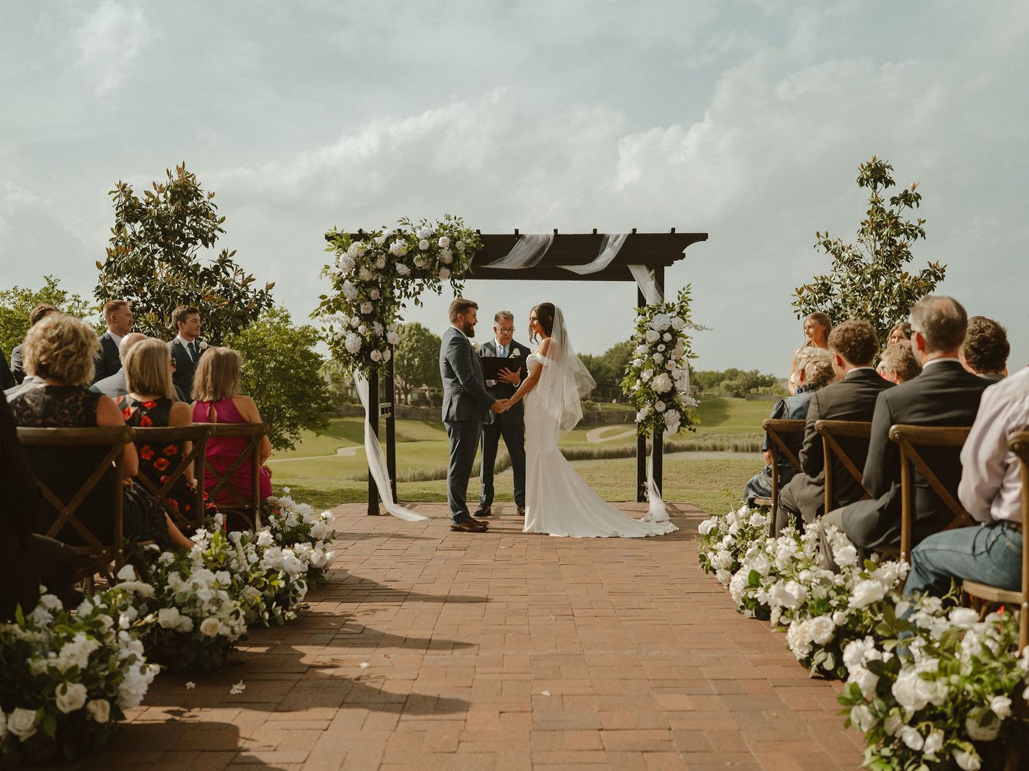 Outdoor wedding ceremony with bride and groom at floral arch, white roses lining brick aisle, guests seated on wooden chairs.