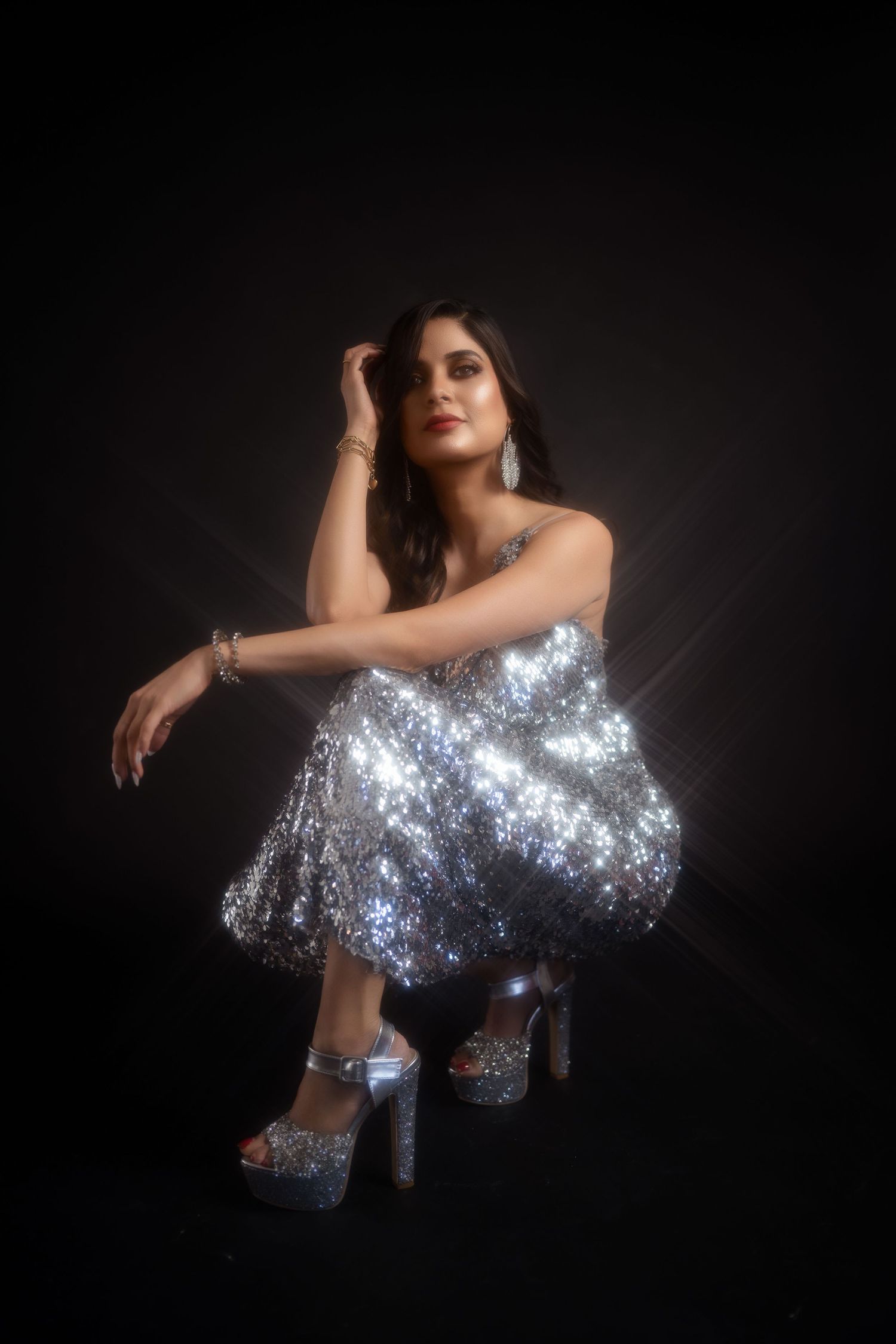 Woman in silver sequin dress and heels posing on dark background with dramatic studio lighting.