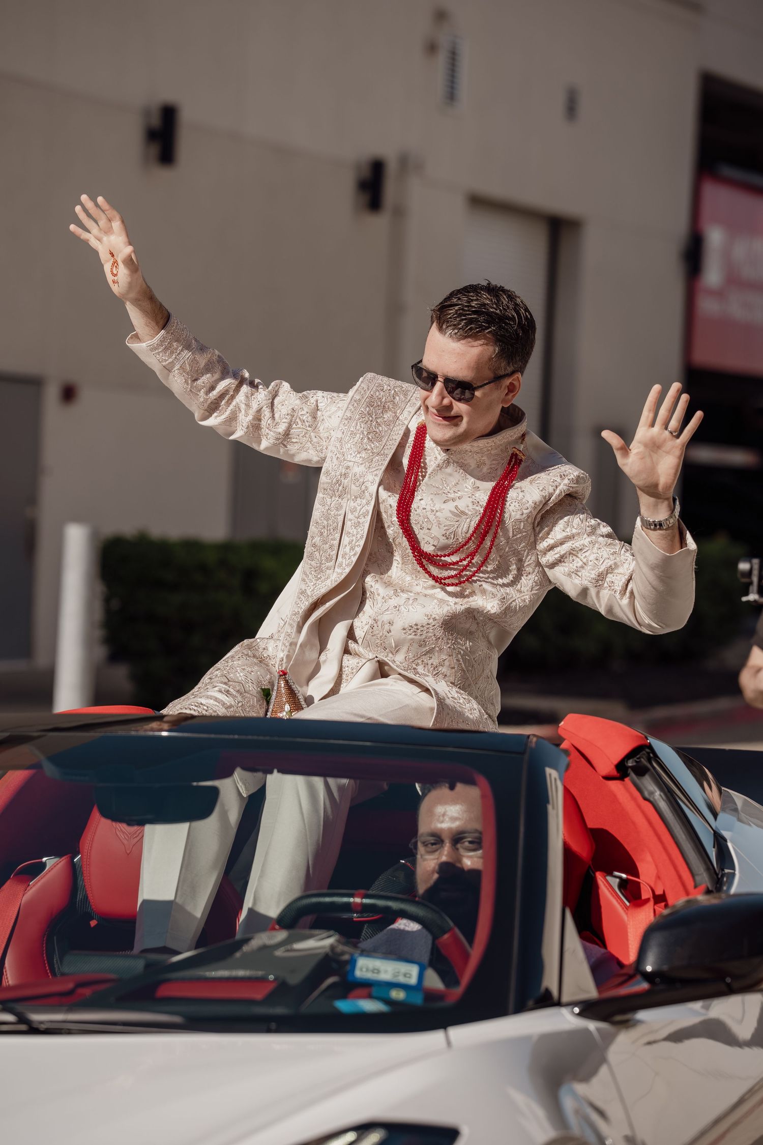 Man in traditional Indian attire and sunglasses standing in a white convertible car with arms raised.