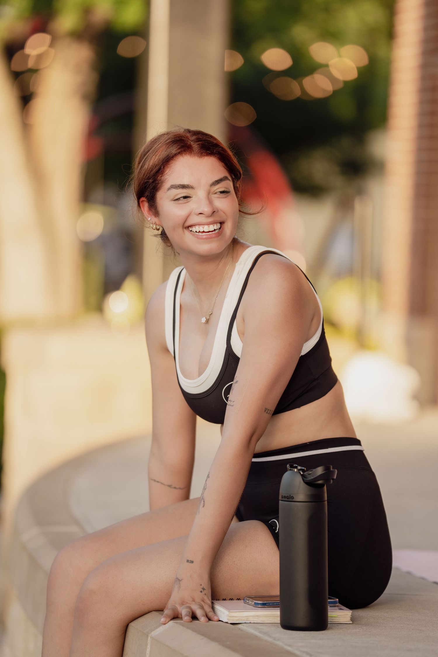 Smiling young woman in black sports bra and leggings sitting outdoors with a water bottle after workout.