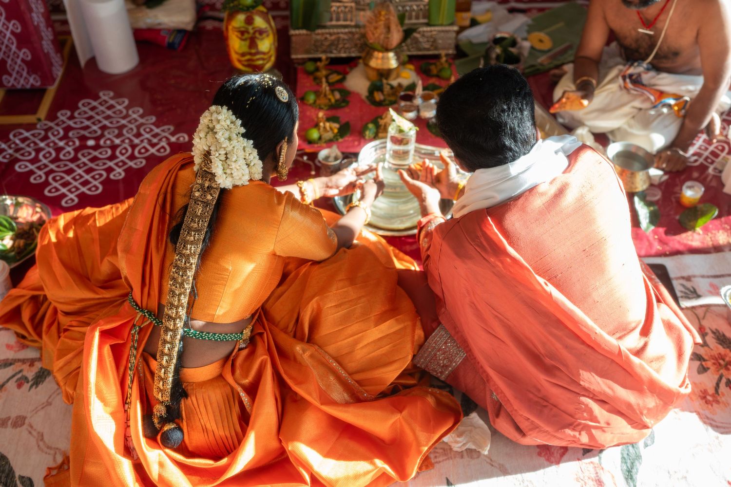 Two people in orange robes performing a traditional Hindu ceremony with ritual offerings and decorations.