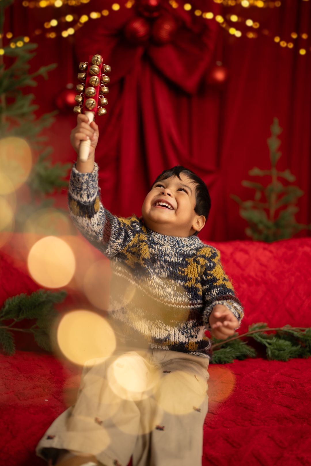 Joyful boy in festive sweater holding jingle bells amid Christmas decorations and bokeh lights.