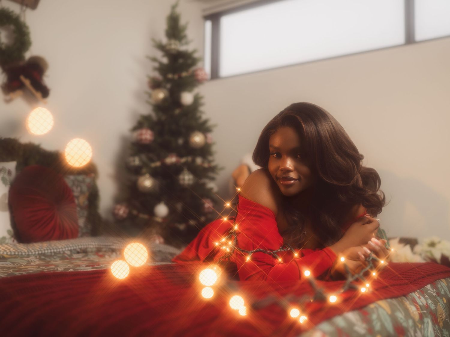 Woman in red dress posing with glowing Christmas lights near a decorated holiday tree indoors.