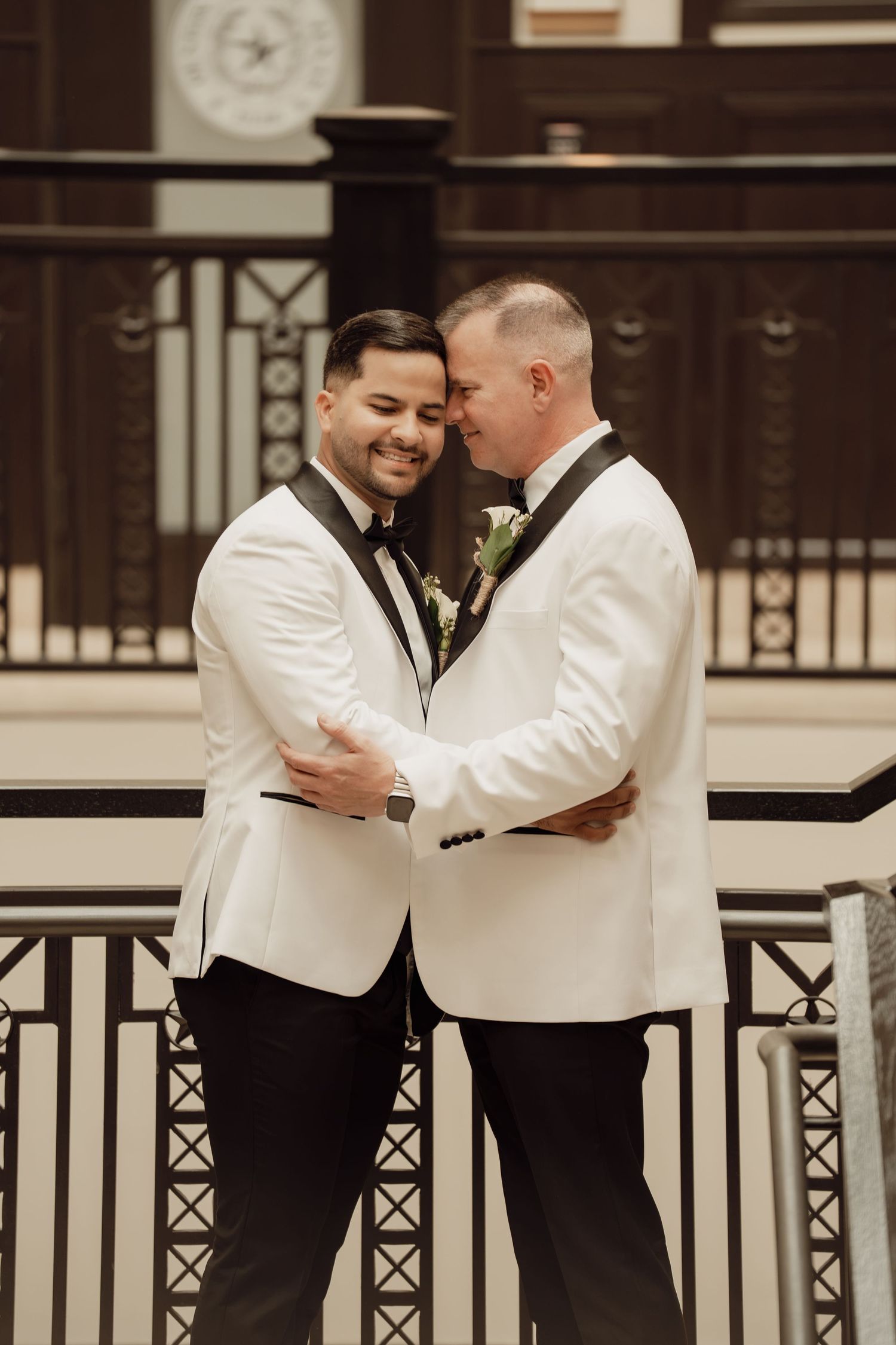 Two grooms in white tuxedos embrace and smile at each other on elegant staircase during wedding ceremony.