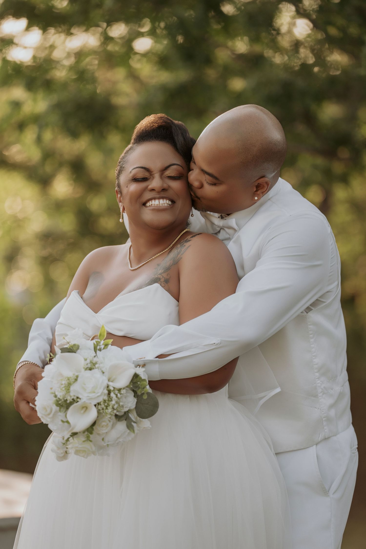 A joyful Black bride in a white gown holds a bouquet as her bald groom in a white suit kisses her cheek outdoors.