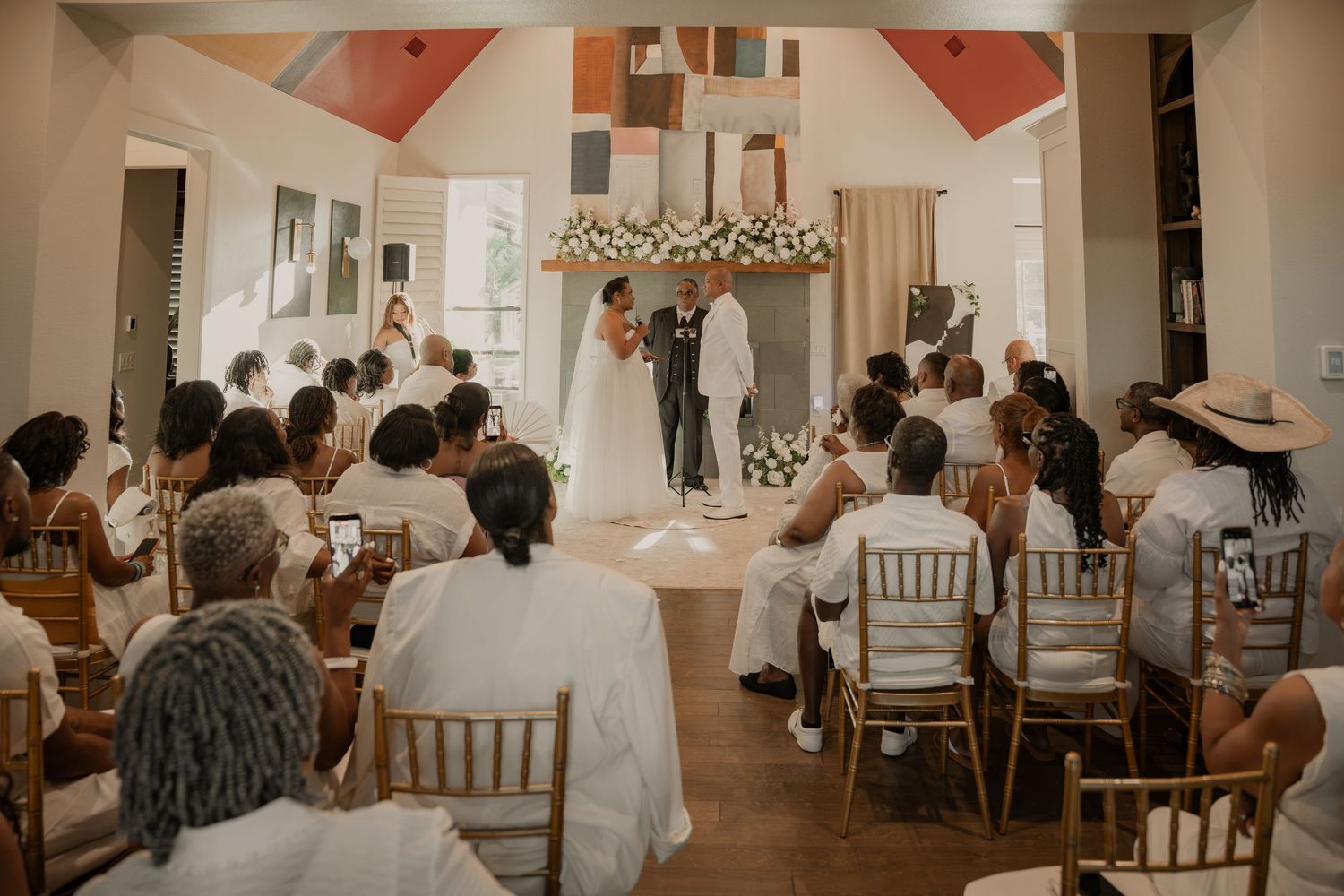 Elegant indoor wedding ceremony with guests seated in gold chairs, couple at floral arch altar in bright white venue.