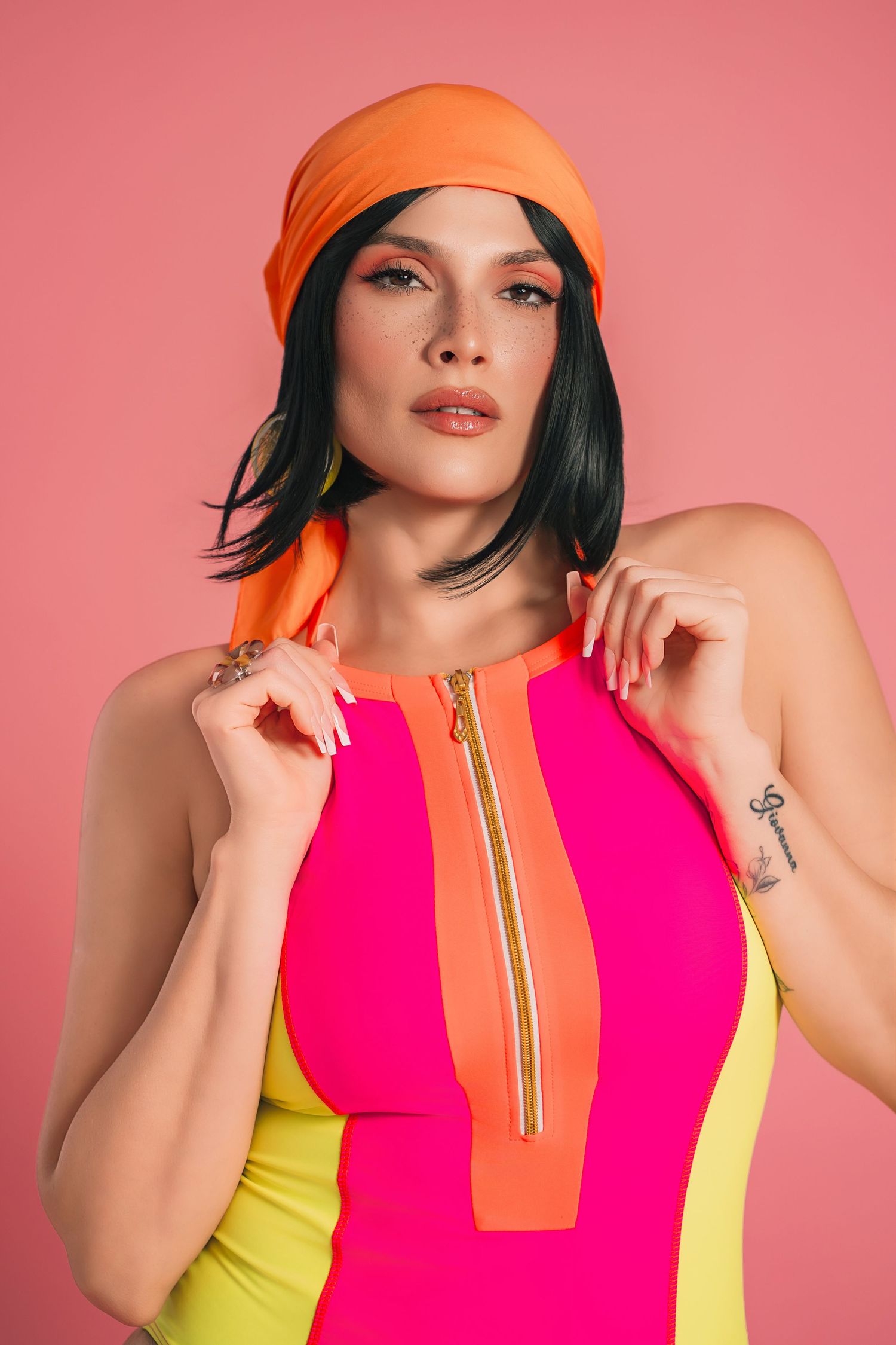 Woman in colorful pink, orange, and yellow zip-up dress with matching orange headscarf on pink background.