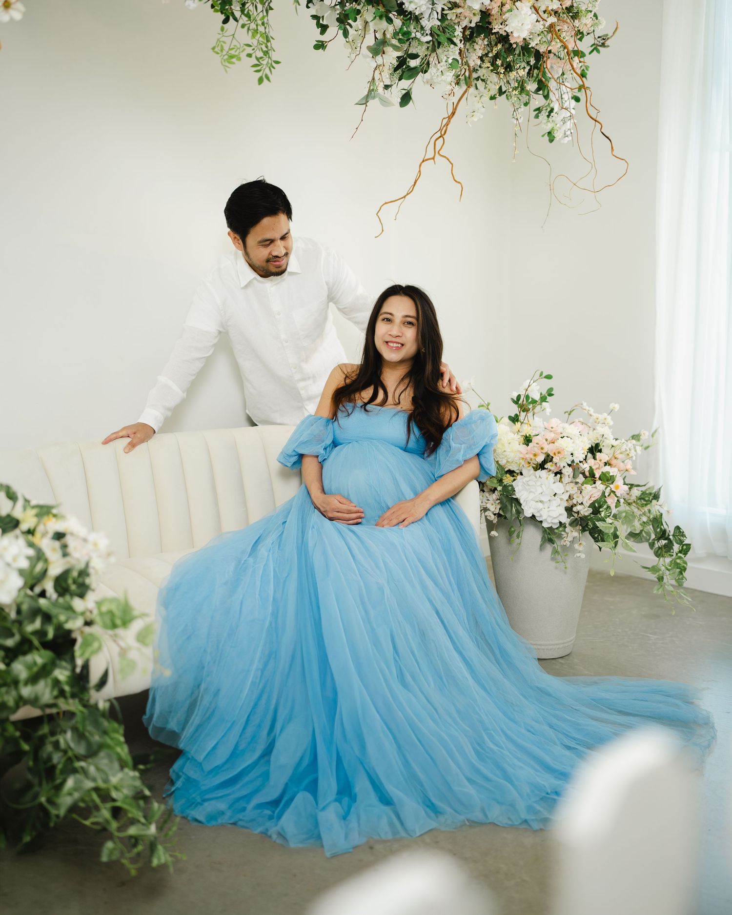 Couple poses for maternity photo shoot, woman in blue tulle gown holding baby bump, man in white shirt, floral decor.