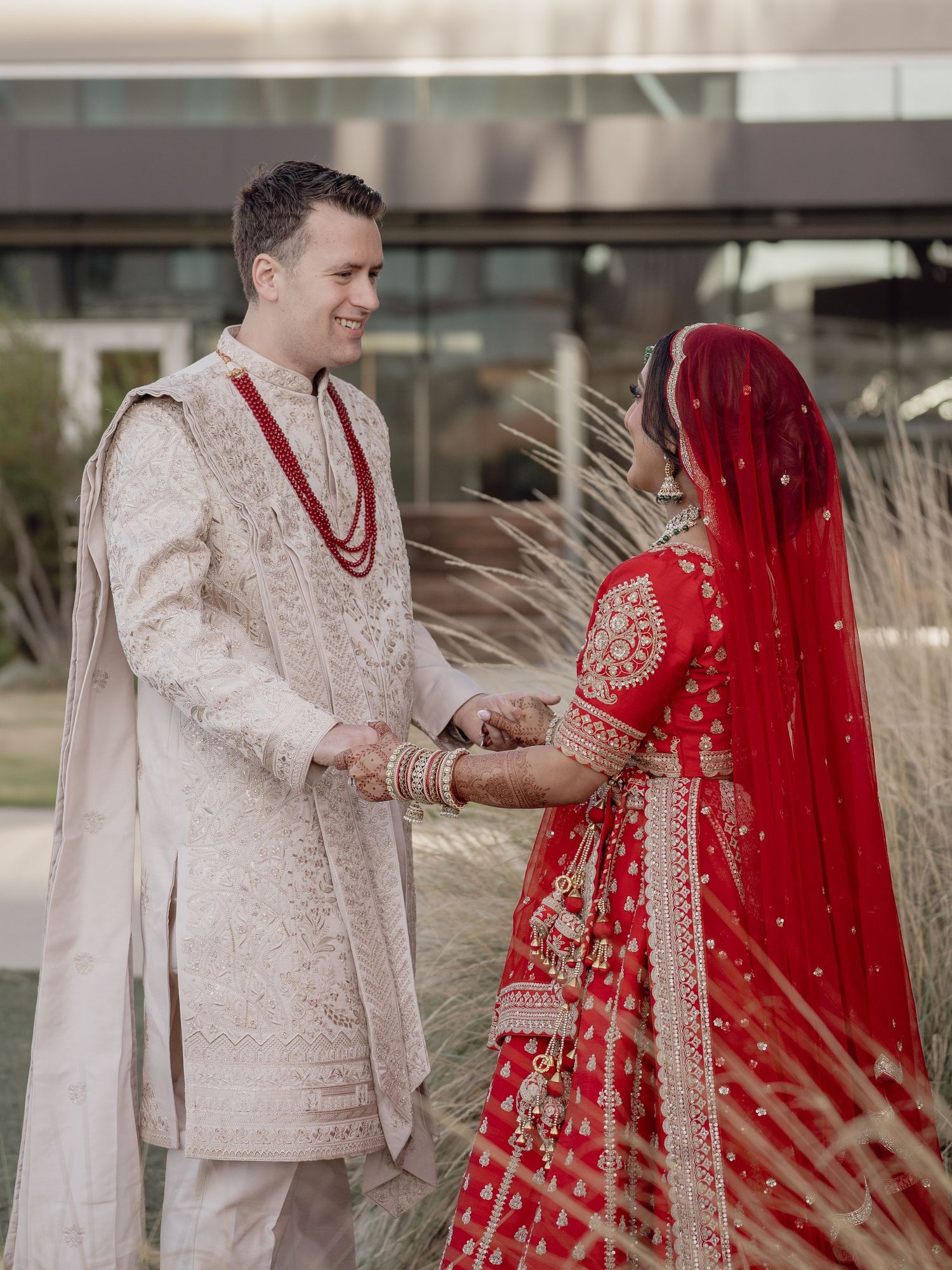 Couple in traditional Indian wedding attire, groom in ivory sherwani, bride in red lehenga with gold embroidery and veil.