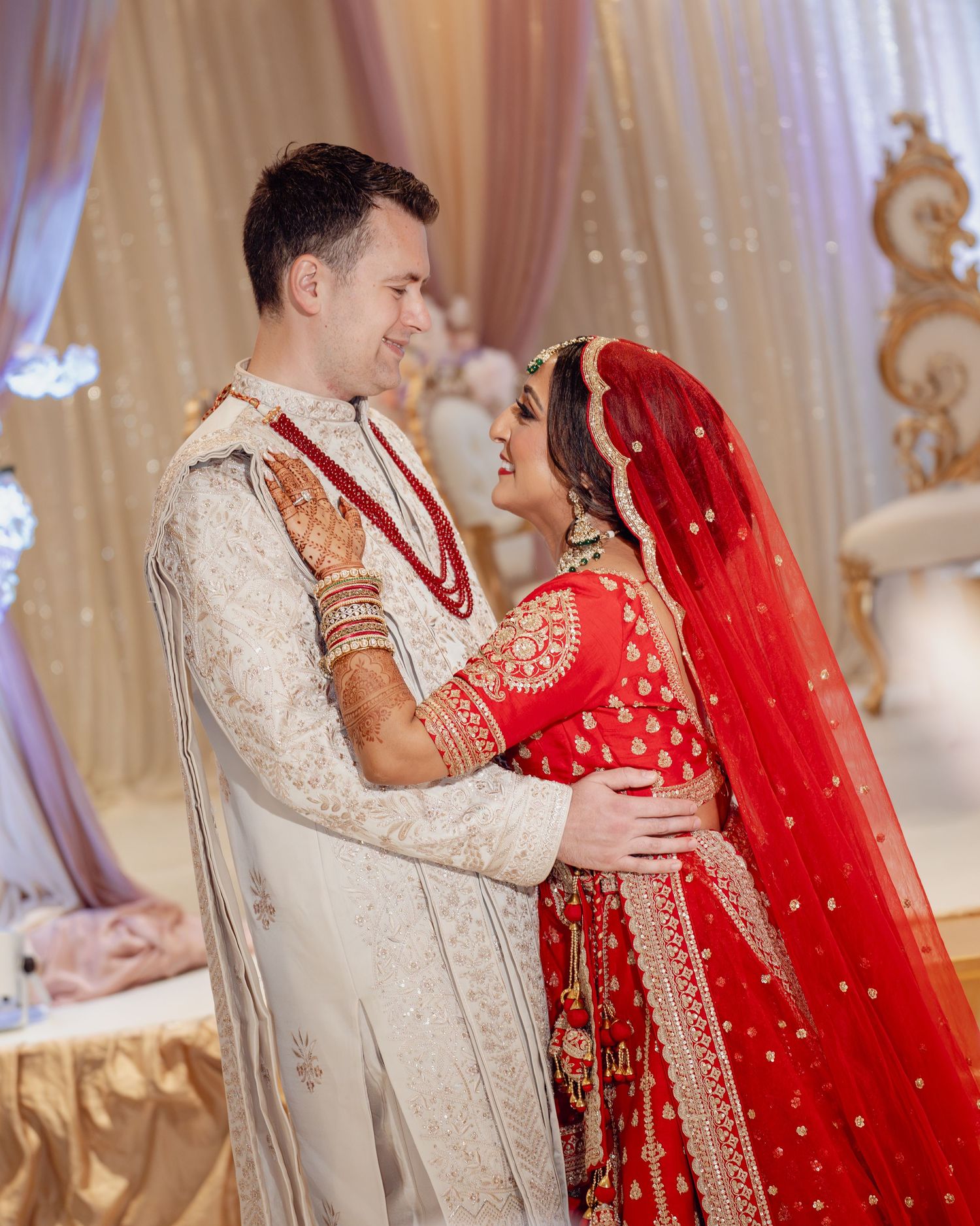 Bride in red lehenga and groom in ivory sherwani share a romantic moment at a South Asian wedding ceremony.