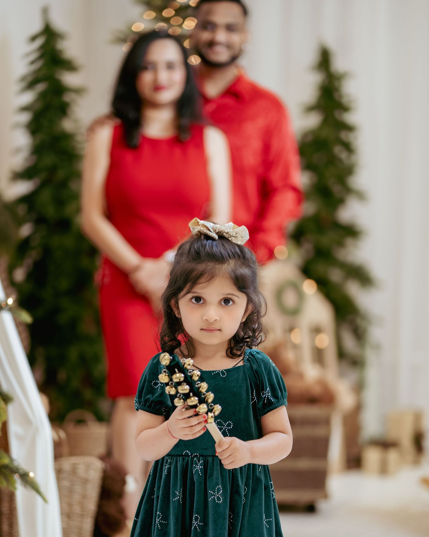 A young girl in a green dress holds holiday bells while her parents in red outfits pose behind her near Christmas trees.