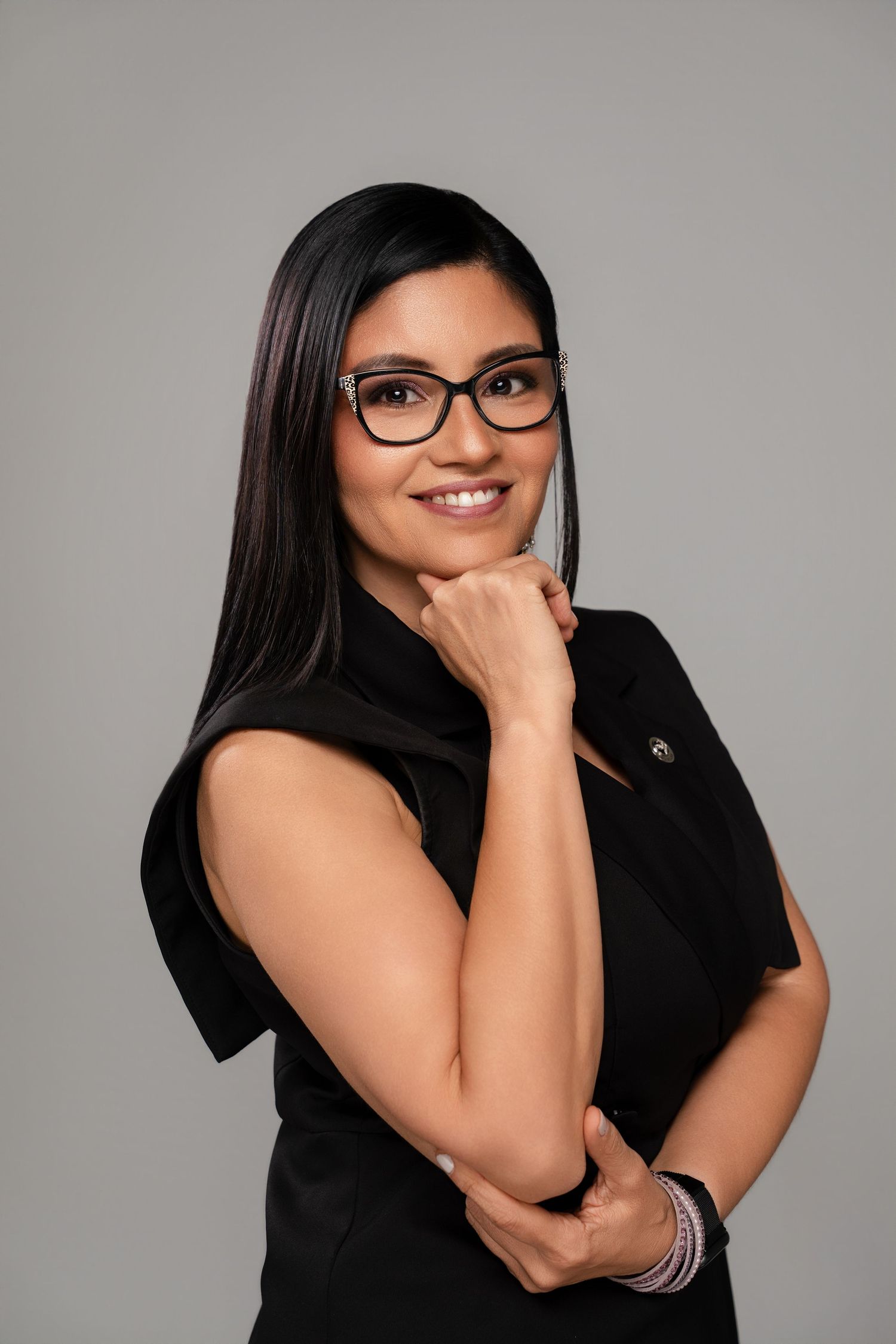 Professional woman in black dress and glasses smiling confidently against gray background.