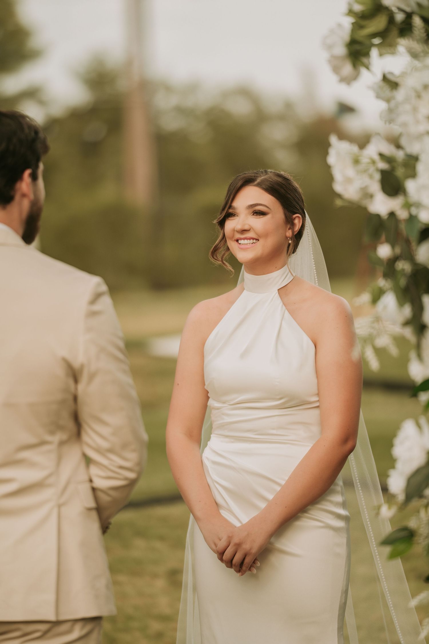 Smiling bride in white halter neck gown and veil stands at outdoor wedding ceremony with white floral arch.