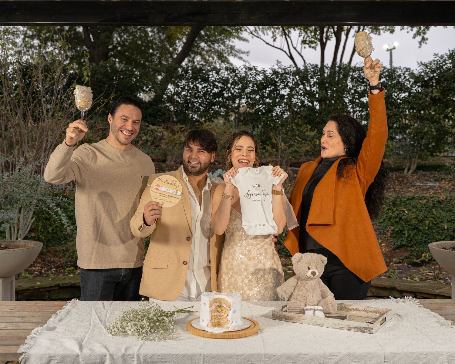 A group celebrates outdoors at a table with a cake and teddy bear while raising their hands in celebration.