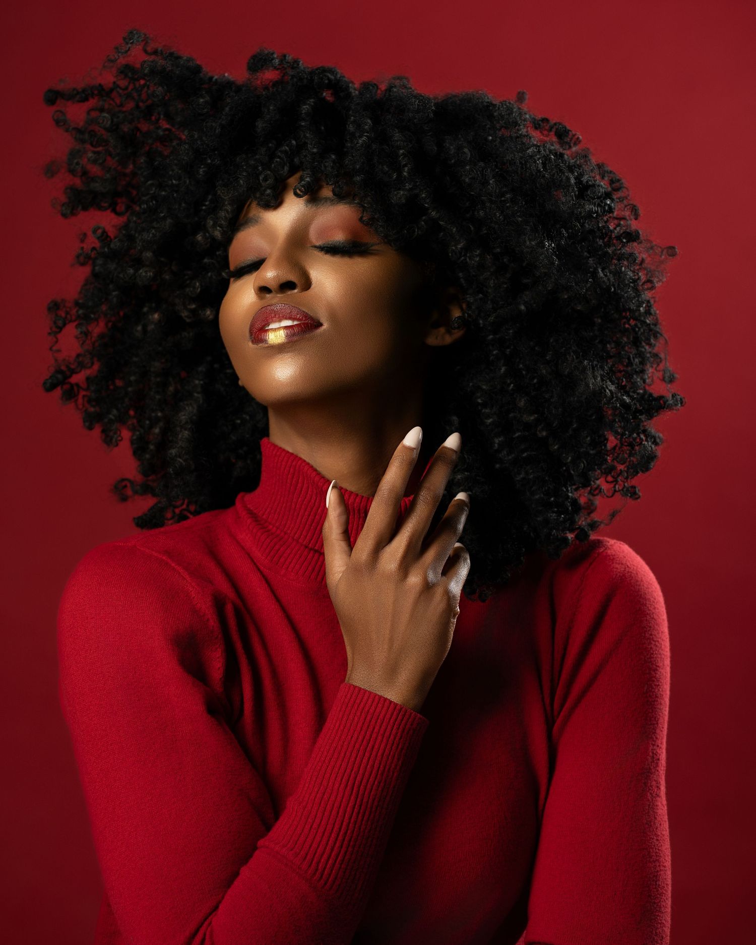 A Black woman with natural curly hair poses confidently in a red turtleneck sweater against a red background.