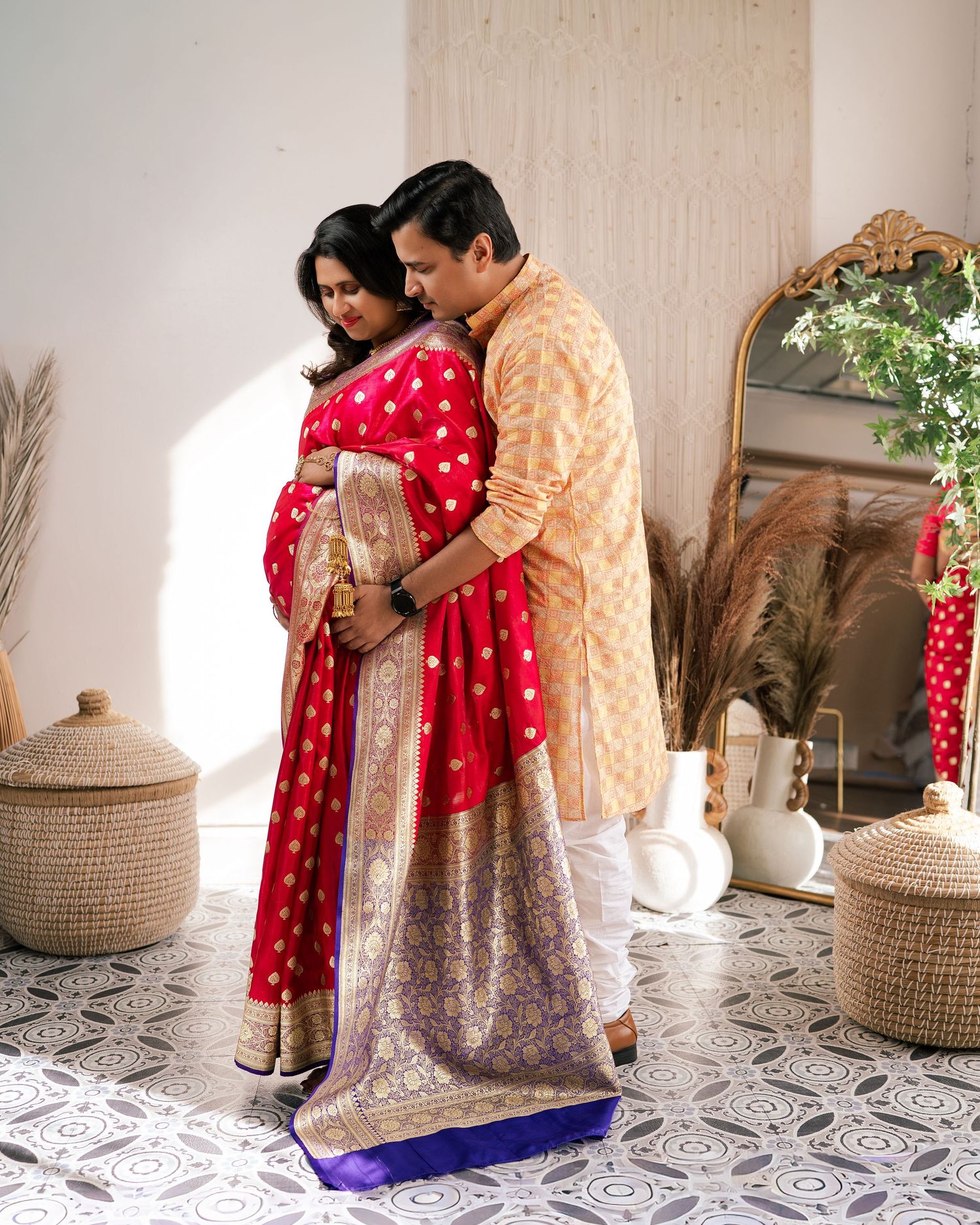 Couple in traditional Indian attire, woman in red saree and man in gold kurta, posing together indoors.