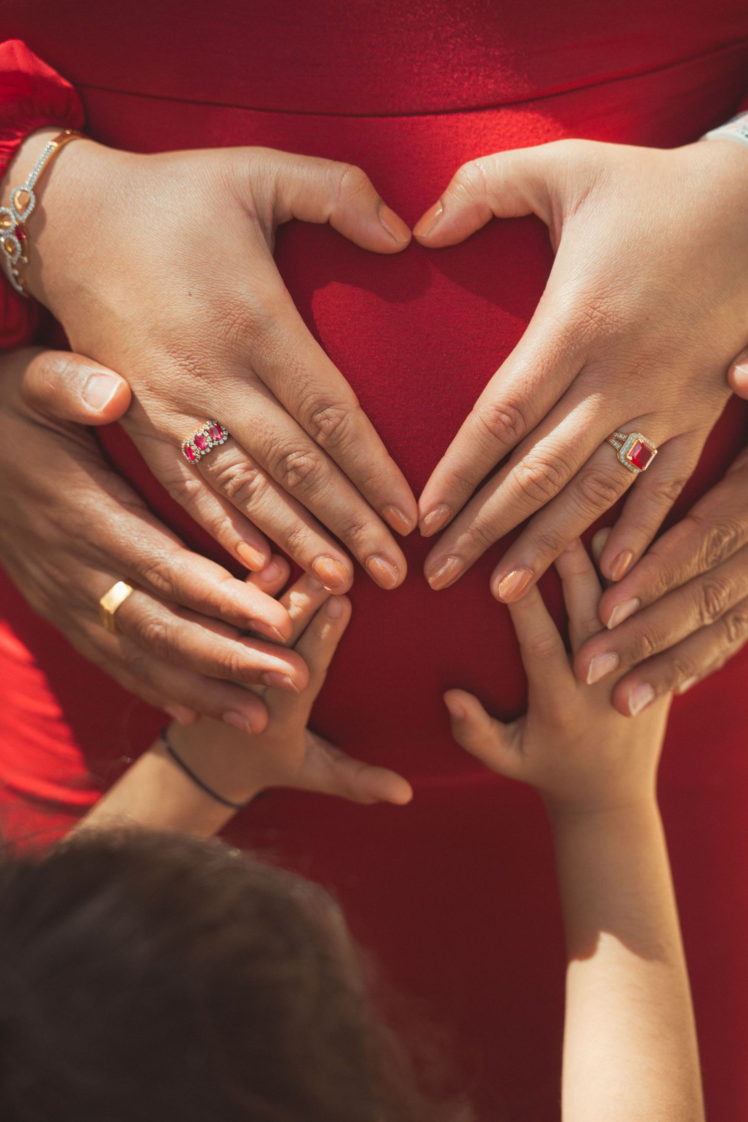 Hands forming a heart shape on a pregnant belly in a red dress, with rings and bracelets visible.