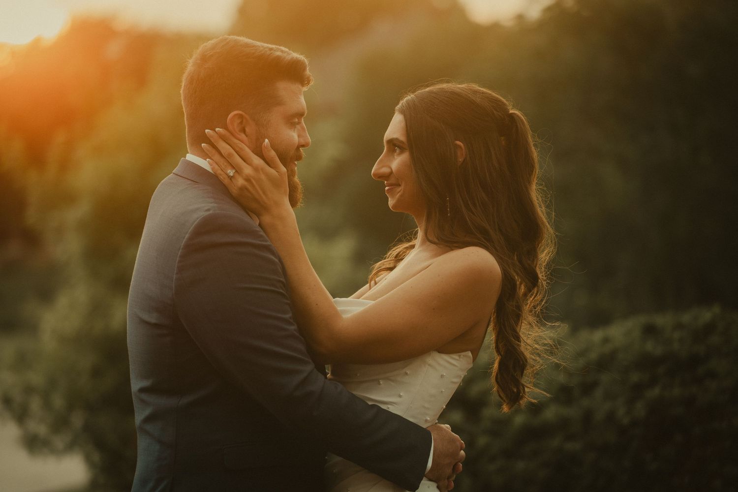 Couple sharing a romantic golden hour moment, bride in white dress with groom in navy suit outdoors.