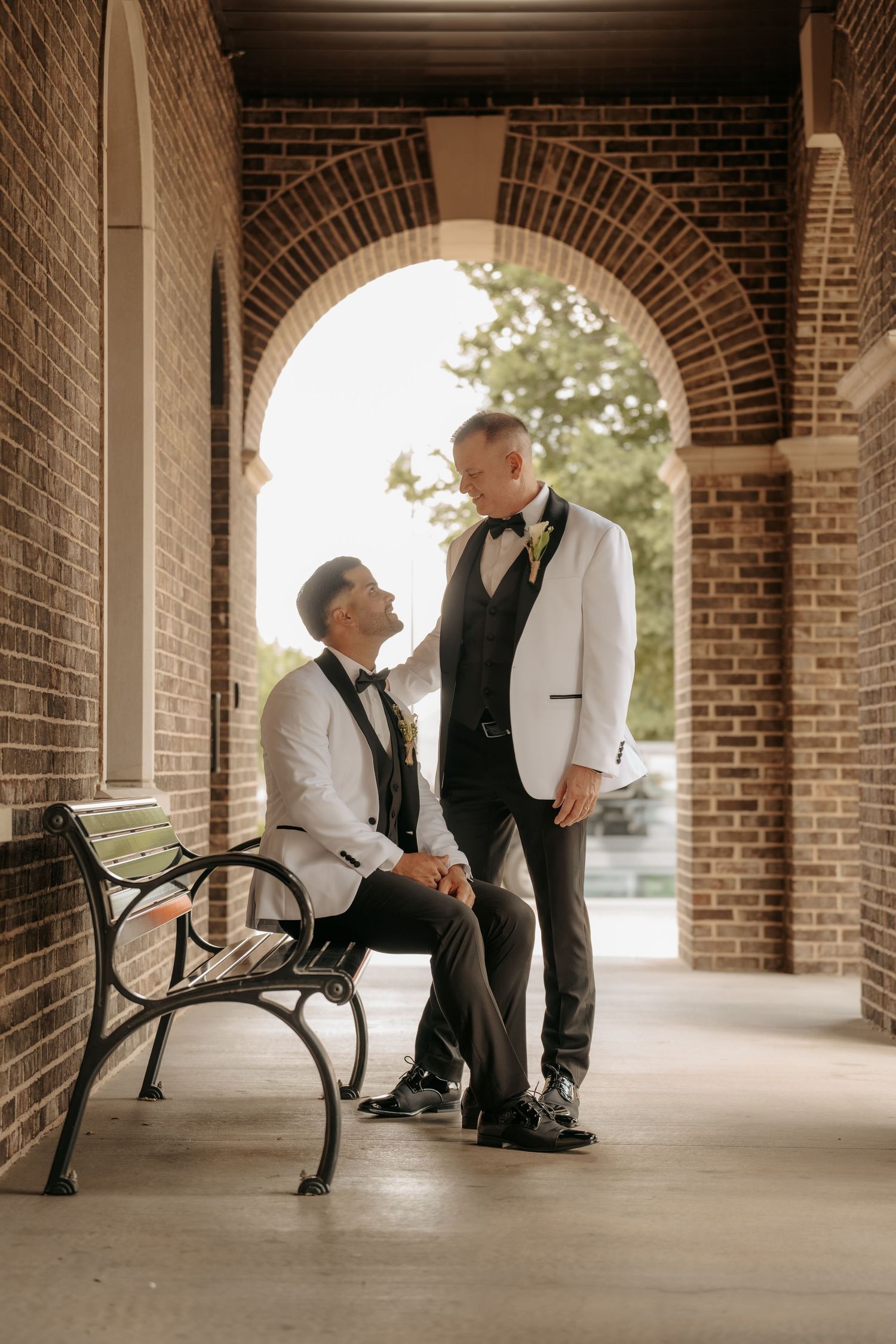Two grooms in white tuxedo jackets share a romantic moment under a brick archway, one seated on a bench.