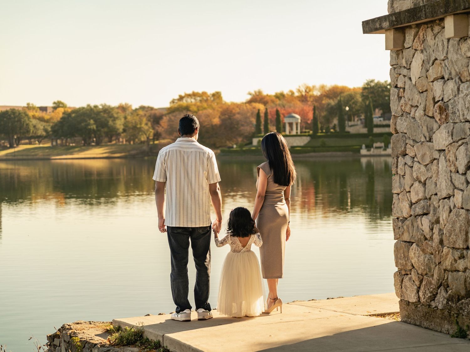 Family of three standing by a serene lake at golden hour, admiring the peaceful water and autumn trees.
