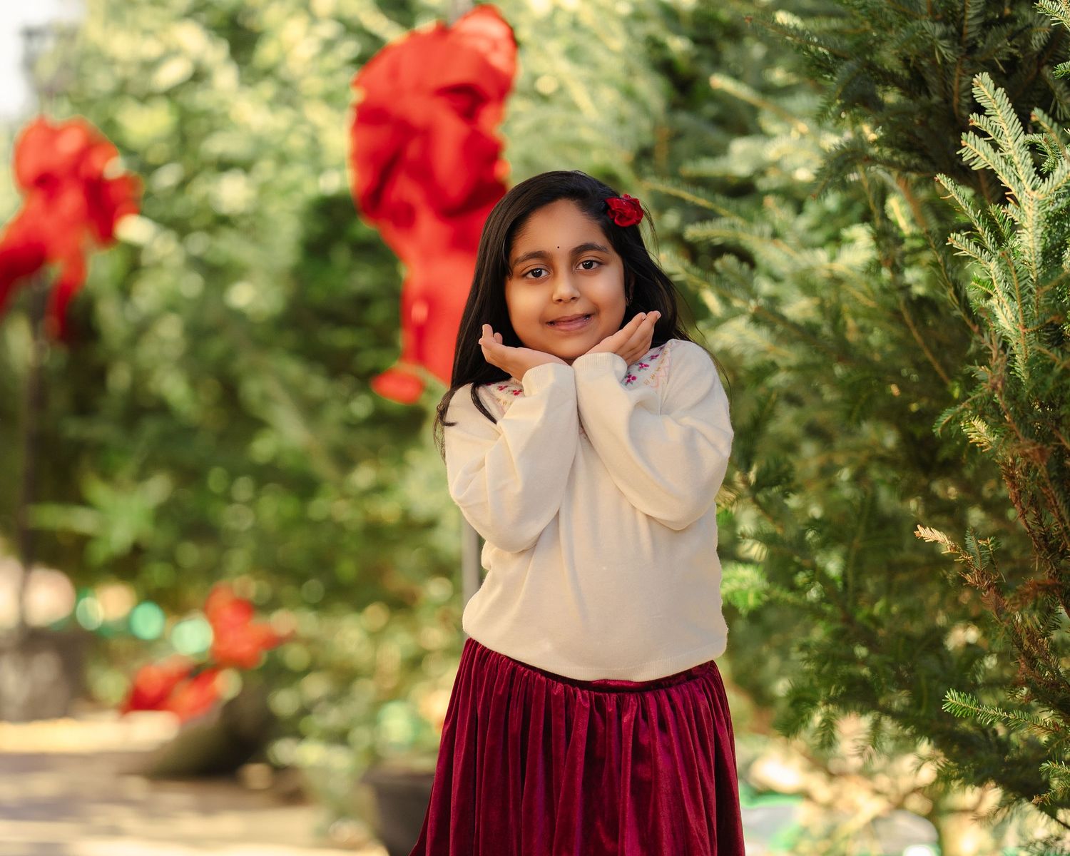 A young girl in a white sweater and red skirt poses among decorated Christmas trees with red bows outdoors.