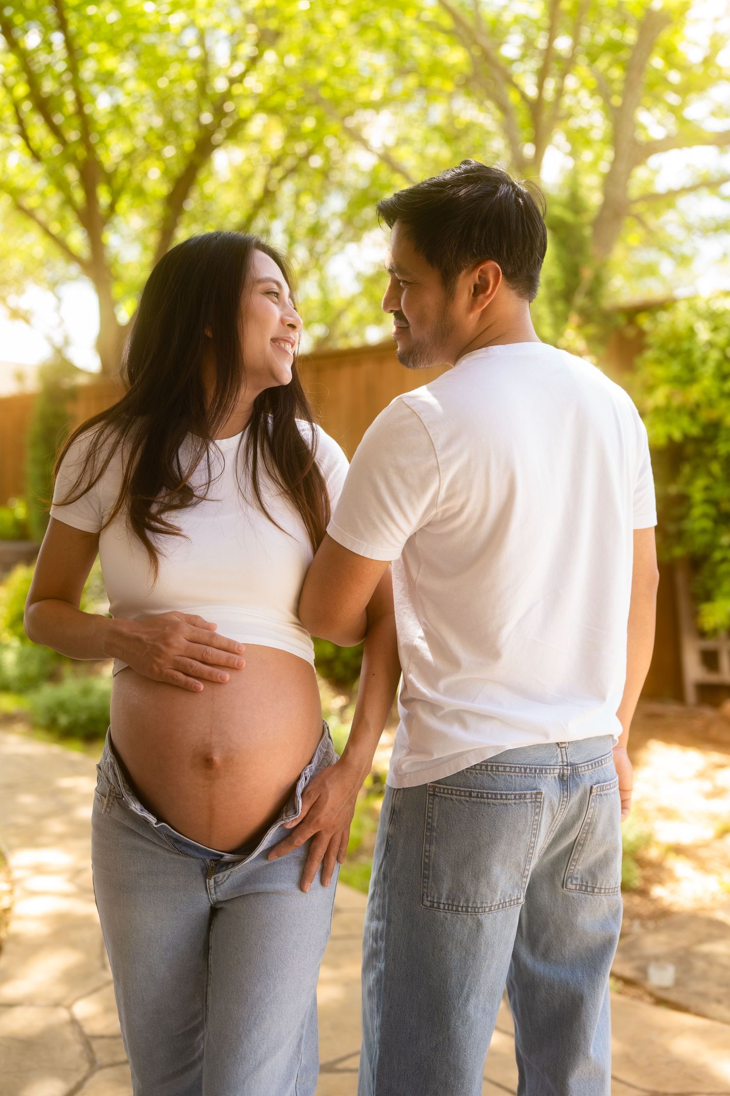 Happy pregnant couple in white shirts and jeans sharing a tender moment outdoors in a sunlit garden.