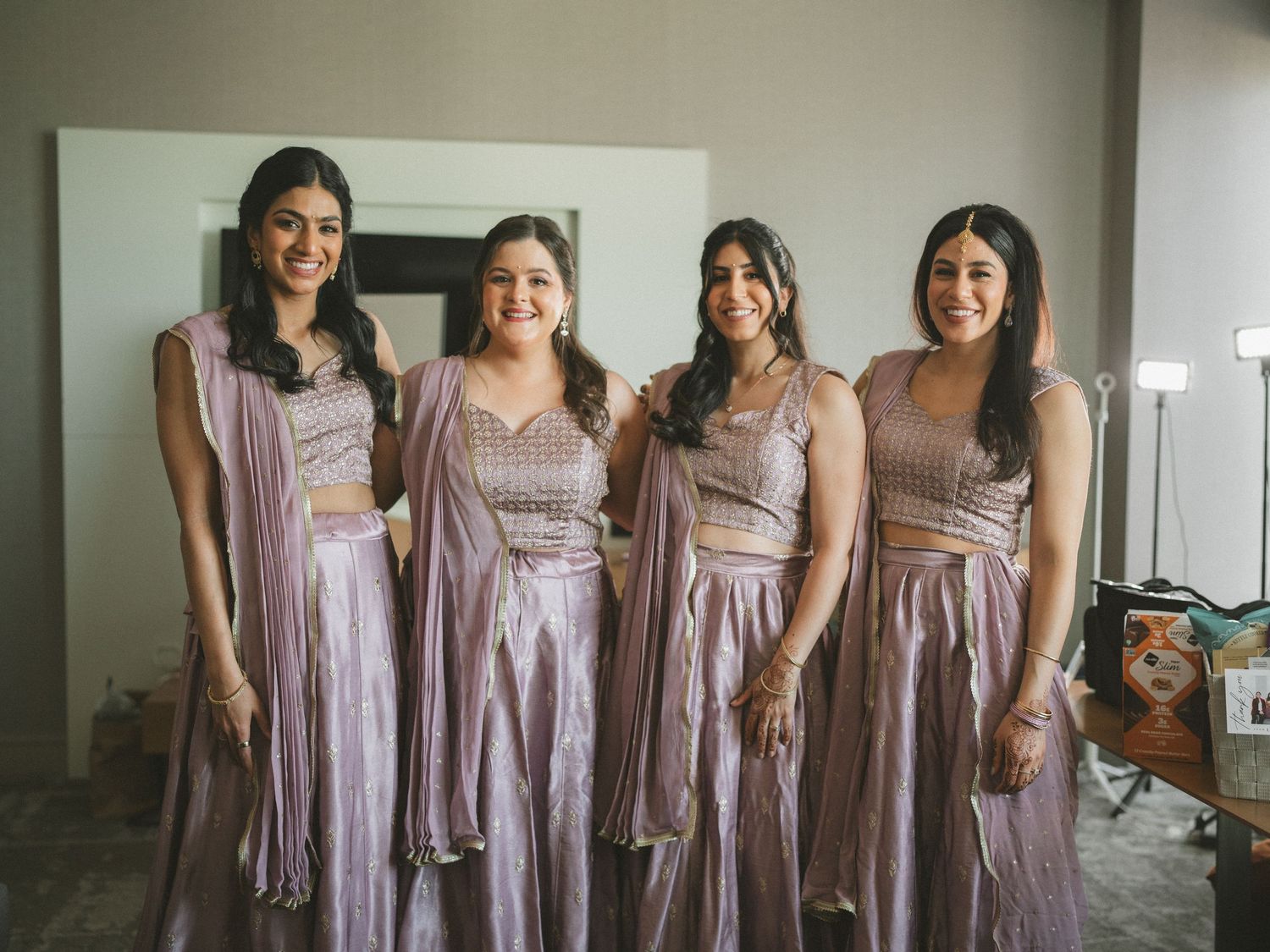 Four bridesmaids in matching dusty rose lehenga outfits smiling together at an Indian wedding celebration.