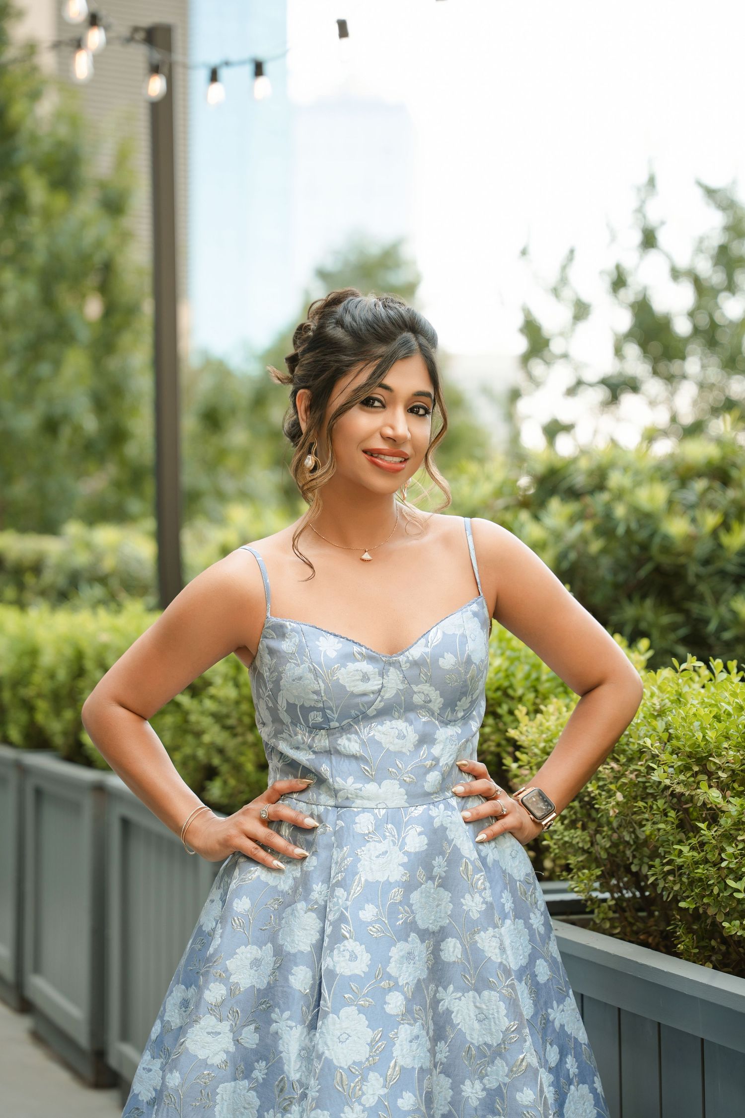 Woman in light blue floral spaghetti strap dress posing with hands on hips outdoors among green shrubs.