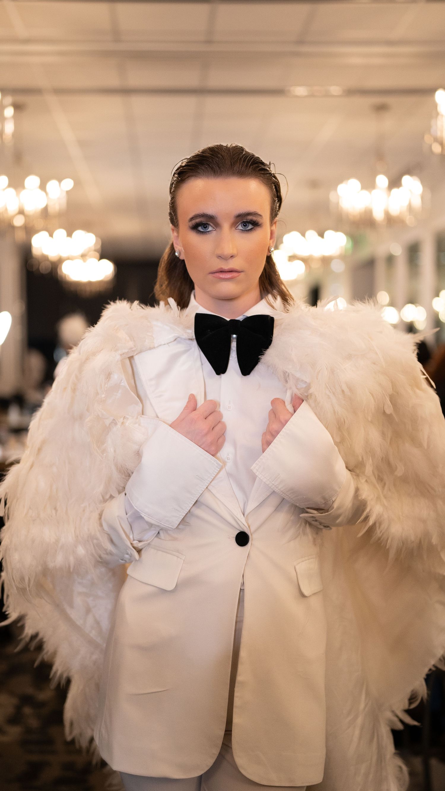 A fashion model wears a white blazer with dramatic feathered angel wings and black bowtie on an elegant runway.