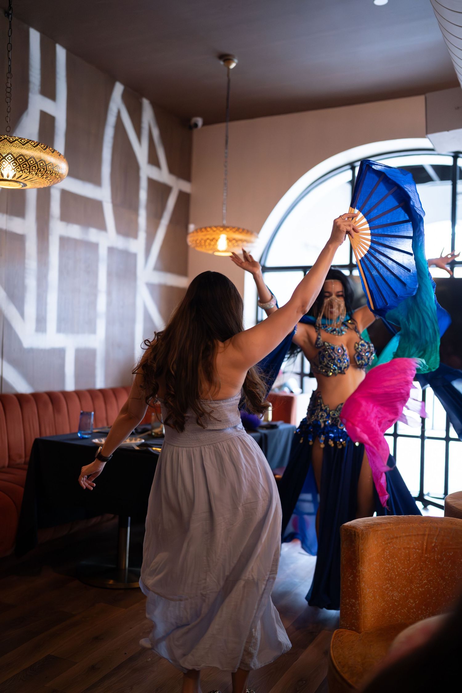 Dancers in flowing costumes perform with blue fans in a dimly lit room with decorative pendant lights.