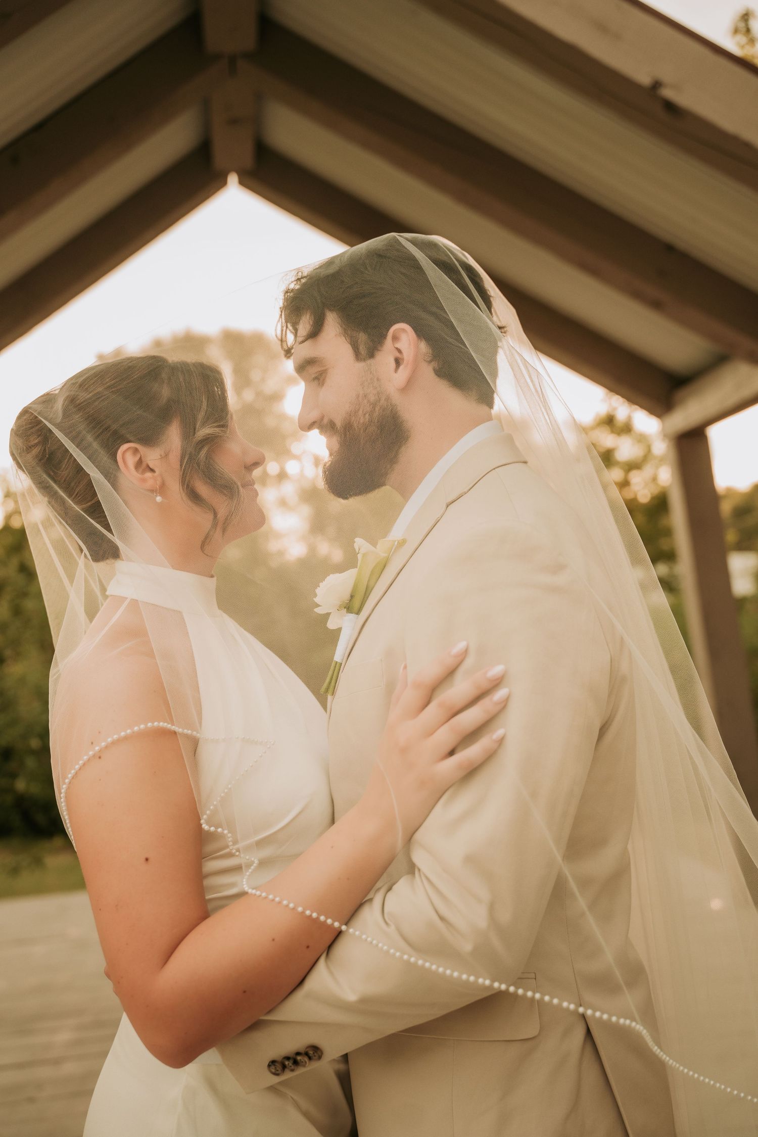 Bride and groom share a romantic moment under a veil at an outdoor wedding venue with warm golden light.