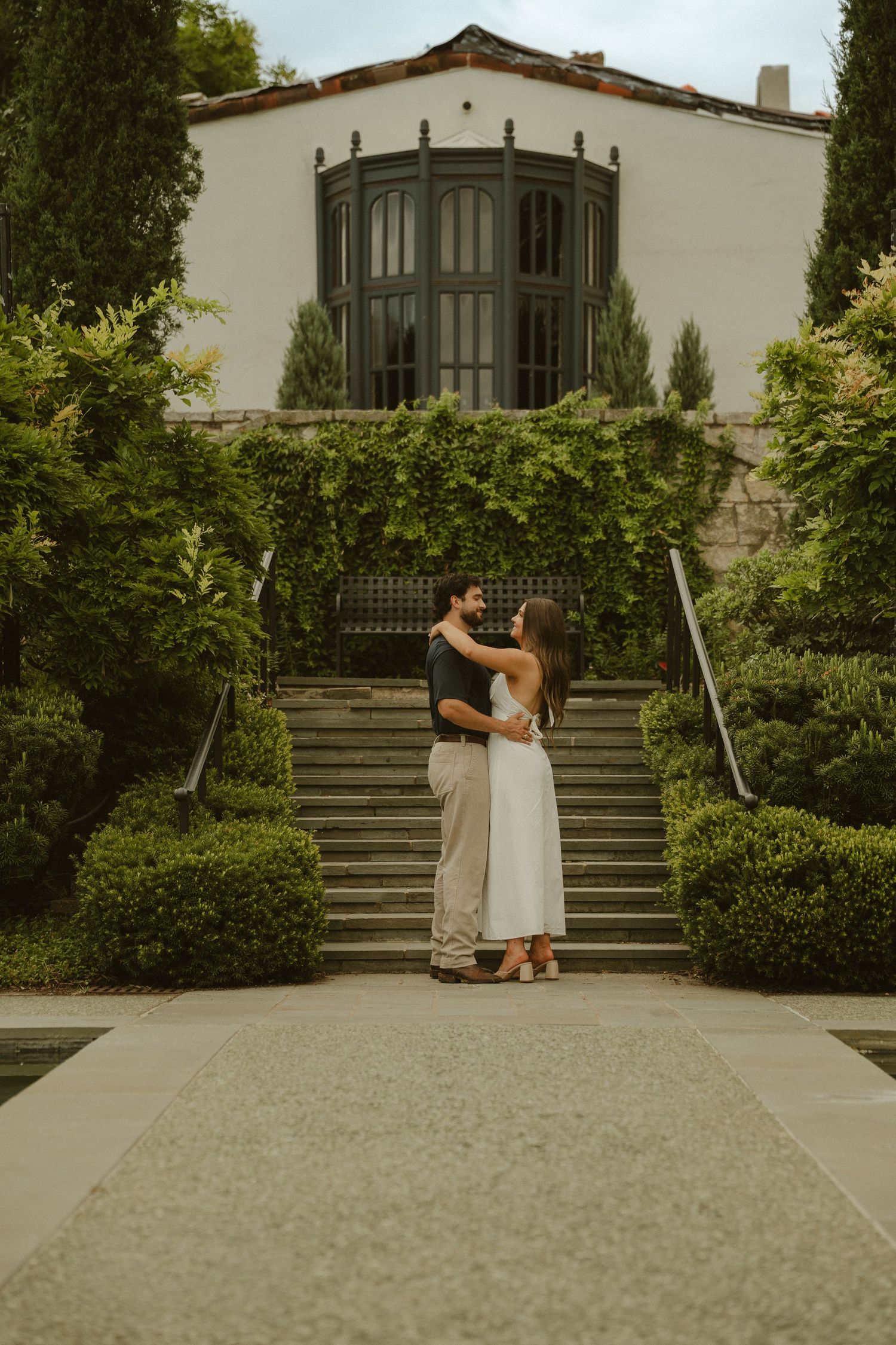 Couple embracing on a garden pathway in front of a white Spanish-style building with lush greenery and stone steps.