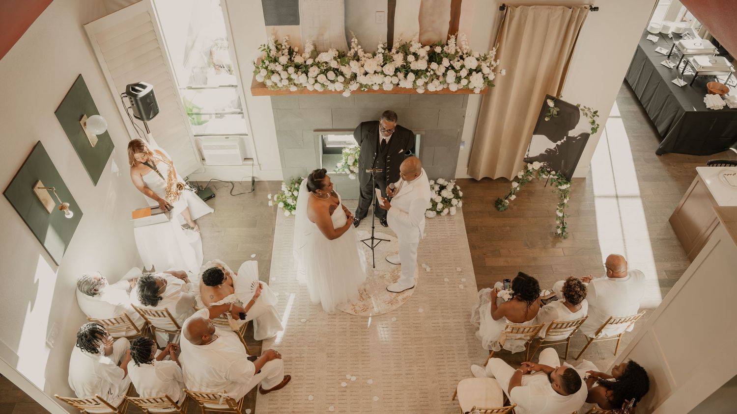 Aerial view of an intimate indoor wedding ceremony with bride and groom at altar, surrounded by seated guests in elegant white decor.