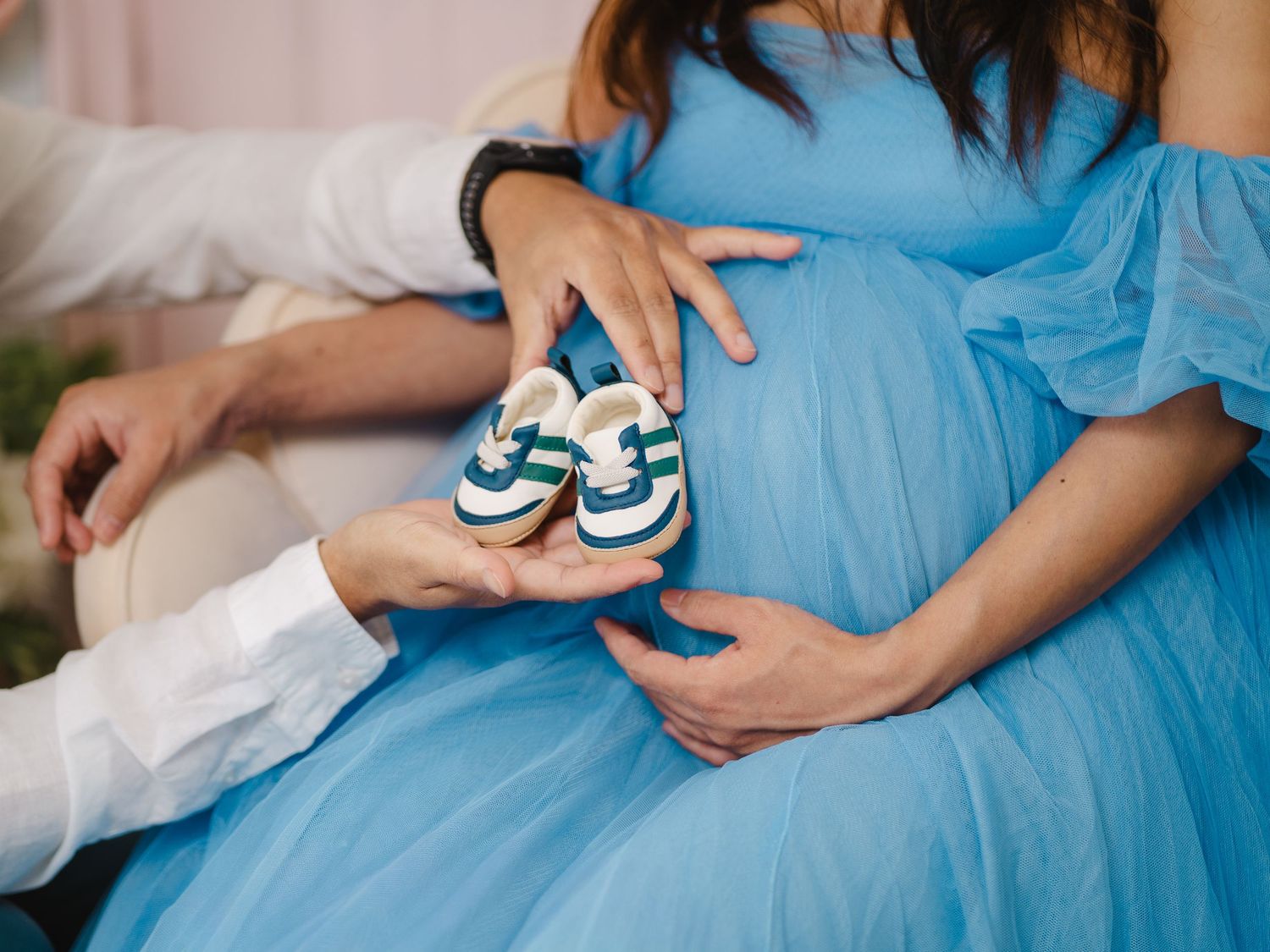 Pregnant woman in blue dress holding tiny baby shoes with partner, maternity photo shoot.
