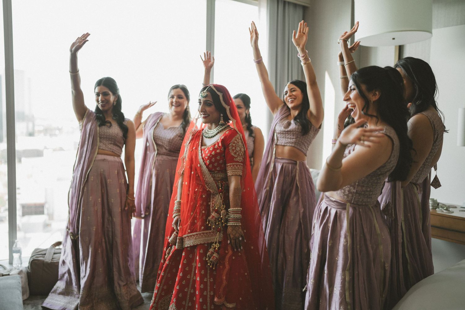 Indian bride in red lehenga celebrates with bridesmaids in mauve outfits, all raising hands joyfully in a bright room.
