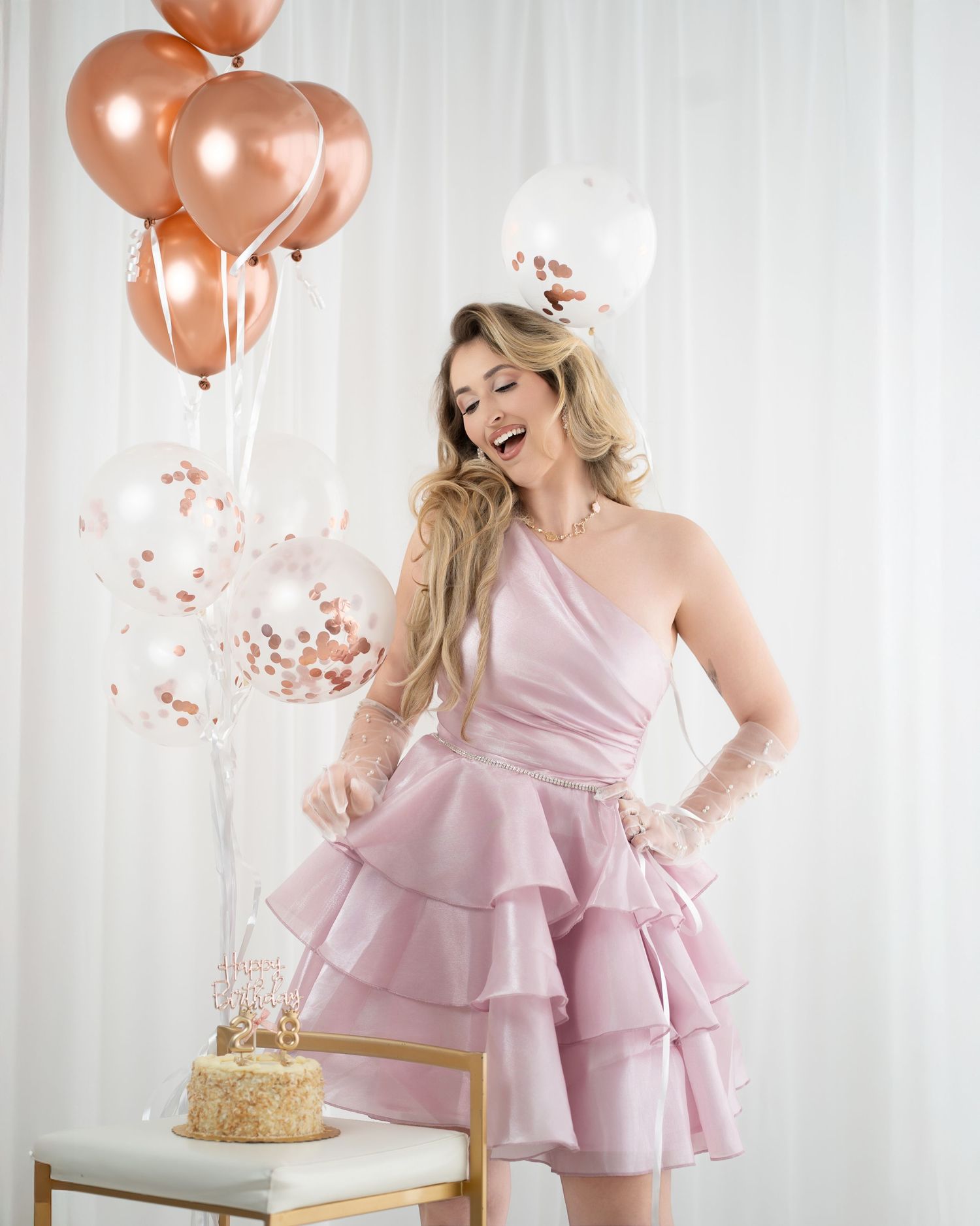 Woman in pink ruffled dress laughing beside rose gold balloons and birthday cake.