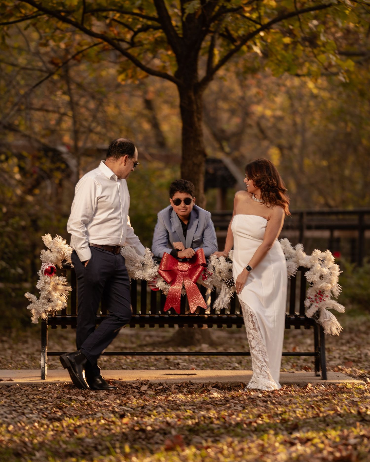 Couple in wedding attire with child on decorated park bench surrounded by autumn foliage.