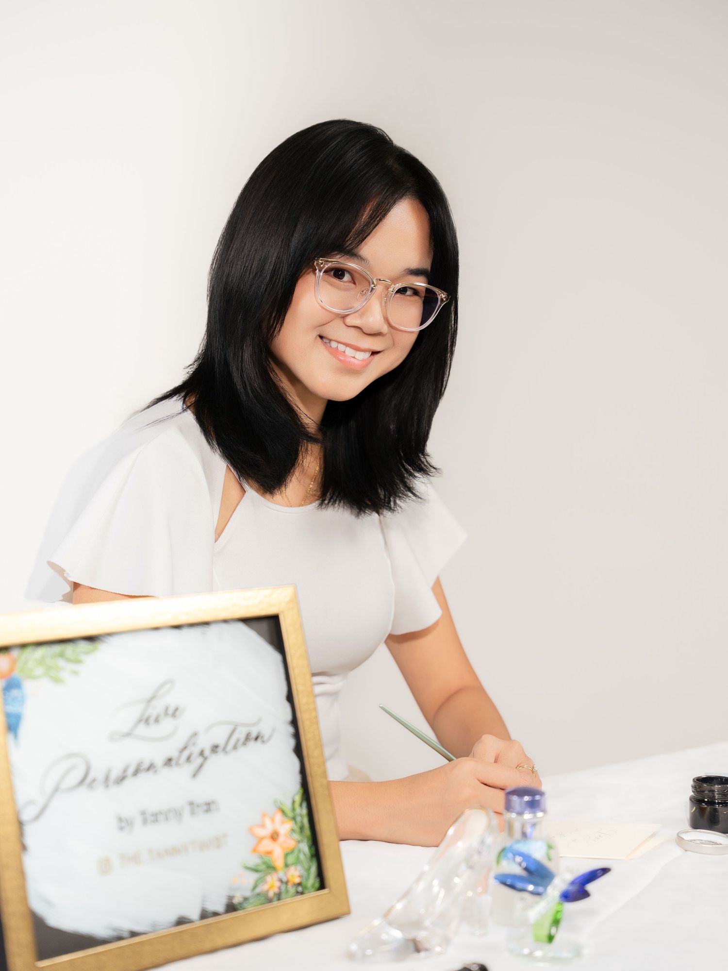 Young woman smiling at a table with a live personalization sign, holding a pen, ready to create custom calligraphy art.