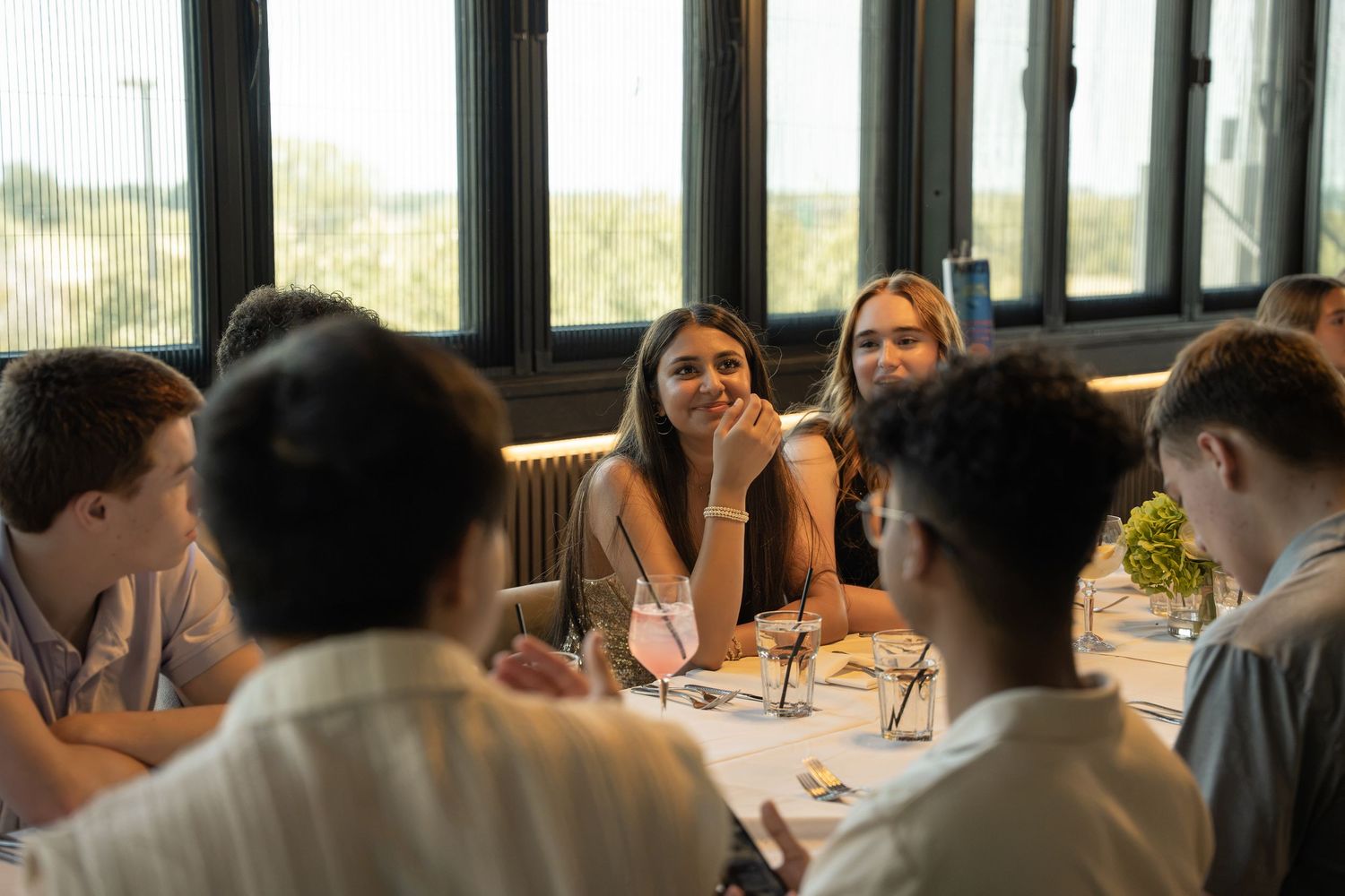 Group of young adults sharing a meal at a restaurant table, engaged in lively conversation near large windows.
