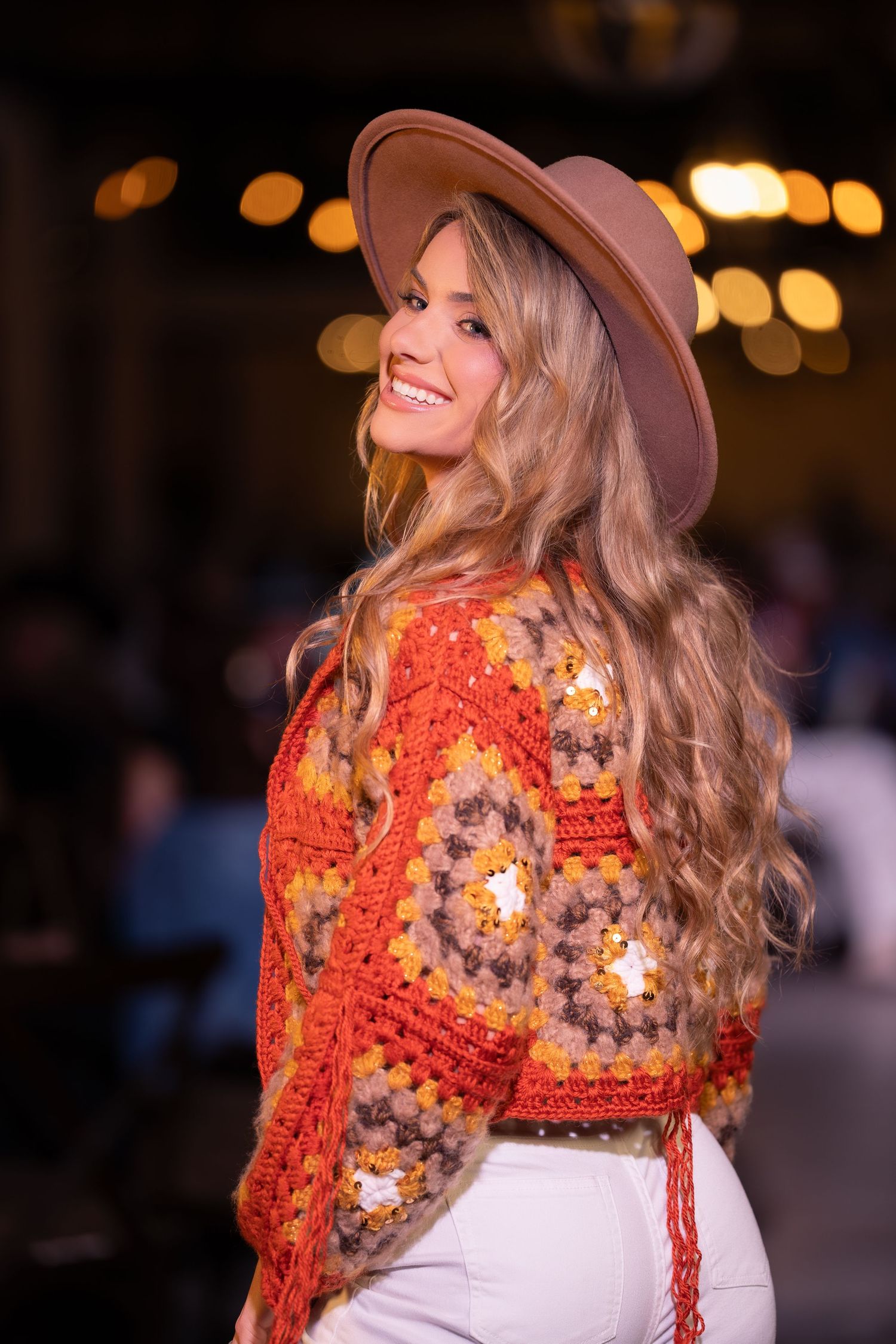 A person wearing a floral orange crochet sweater and tan hat smiles while posing against blurred evening lights.