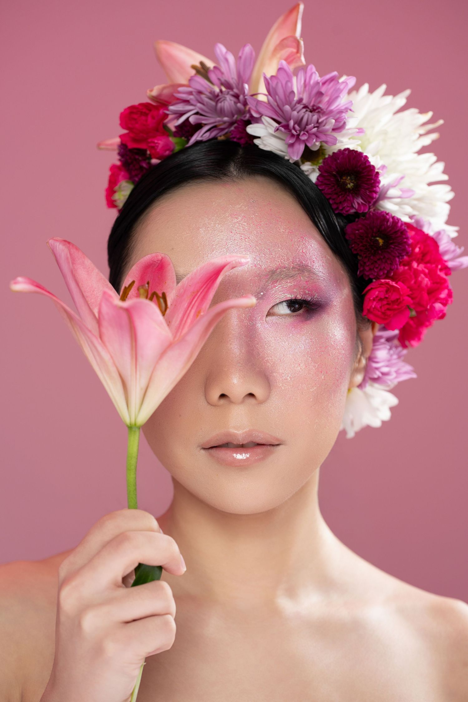Asian woman with floral crown holding pink lily flower near face against pink background.