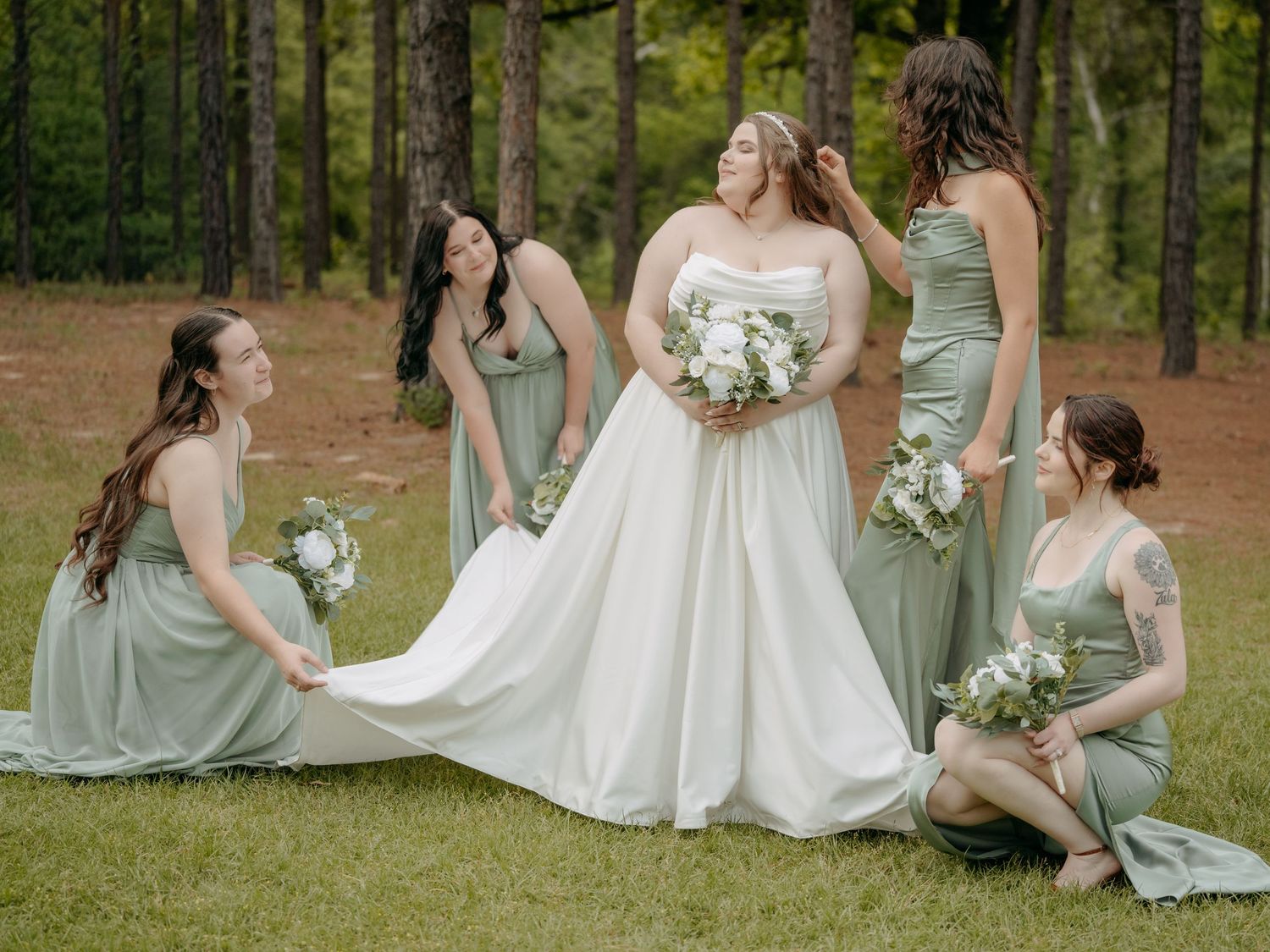 Bride in white gown with bridesmaids in sage green dresses posing outdoors in a pine forest setting.